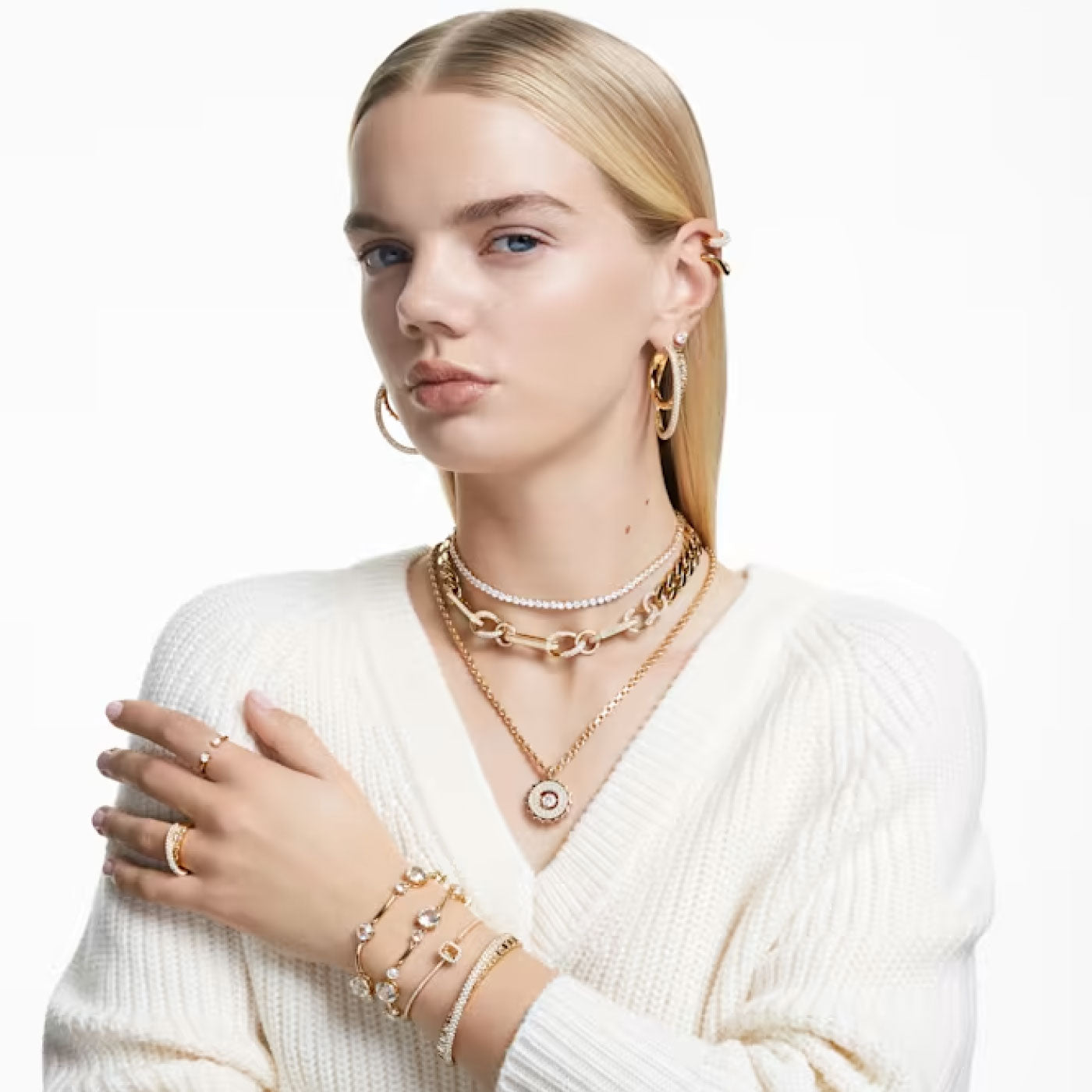 A person with long, blond hair is wearing a white sweater adorned with multiple gold-tone plated necklaces. Hoop earrings, bracelets, and the Swarovski Mixed Cuts Rota Cocktail Ring set with baguette cut stones complement their look. With a confident expression, they are posed against a plain white background.