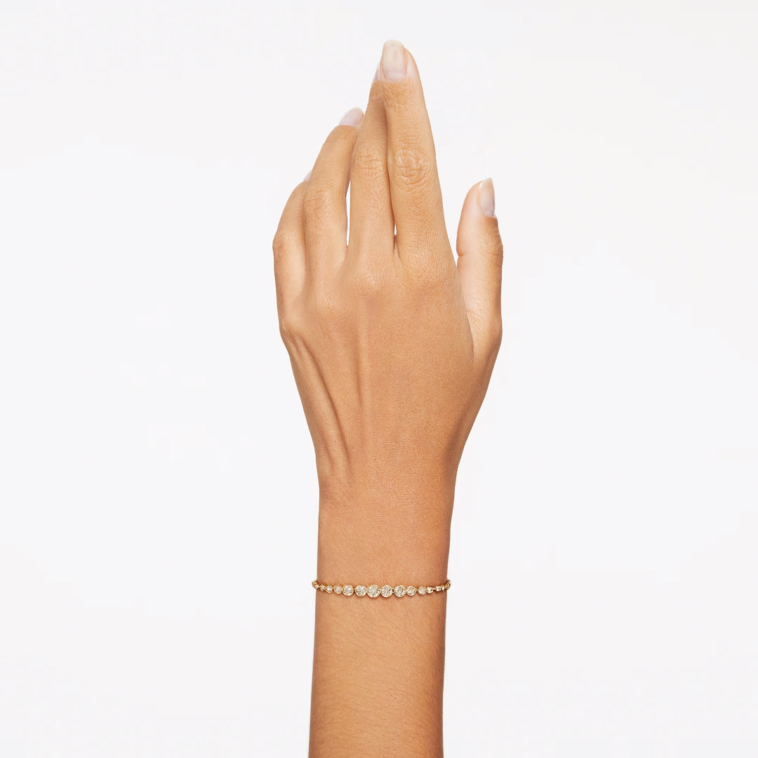 A hand wearing the elegant Swarovski Imber Emily Bracelet, featuring a delicate gold-tone with evenly spaced small white stones, is held up against a plain white background. The sliding clasp adds functionality, while the bracelet's simplicity and elegance are beautifully highlighted by the uncluttered setting.