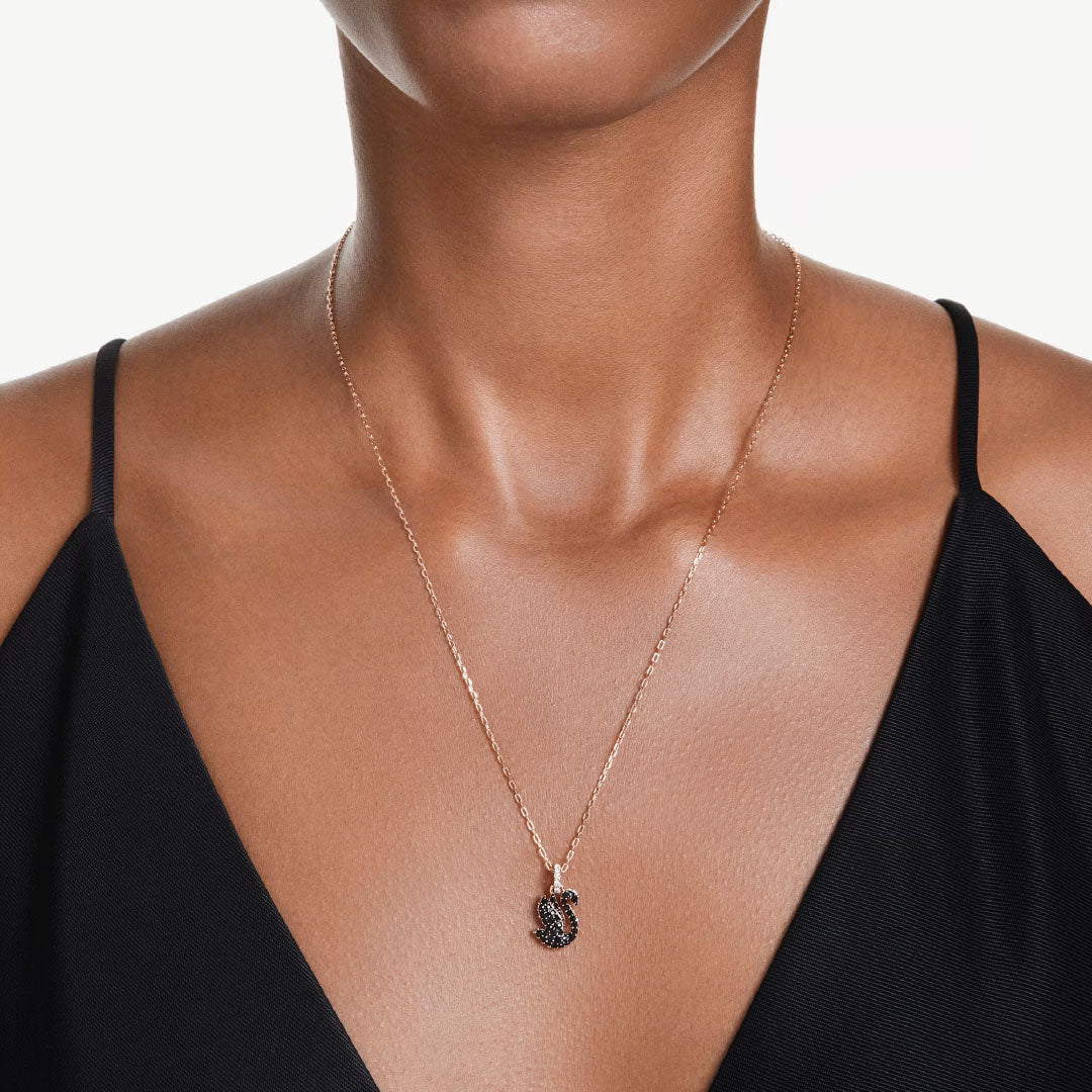 Wearing a black sleeveless top, a person showcases the Swarovski Swan Necklace Pendant by Swarovski, featuring a captivating pendant adorned with ReCreated crystals in an intricate design that stands out against the simple backdrop.