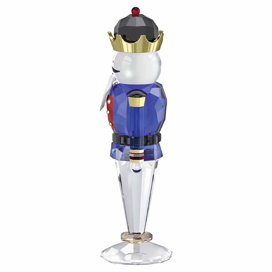 The Swarovski Holiday Cheers Blue Nutcracker Home Décor by Swarovski showcases a toy soldier figurine reminiscent of a nutcracker, adorned in a blue uniform and gold crown. Enhanced with colorful accents, it stands gracefully on a clear base that brilliantly reflects light, making it an ideal holiday decoration.