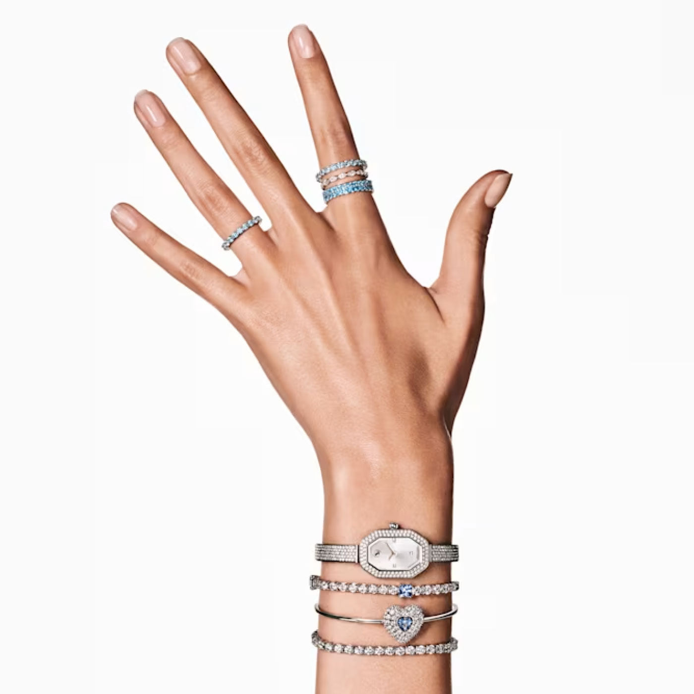 A hand decorated with silver and diamond bracelets and rings showcases a Swarovski Hyperbola Heart Bangle encrusted with Swarovski Zirconia. The wrist is complemented by an elegant watch, while the fingers feature stylish rings, including one with a heart-shaped dancing stone, all set against a plain white background.