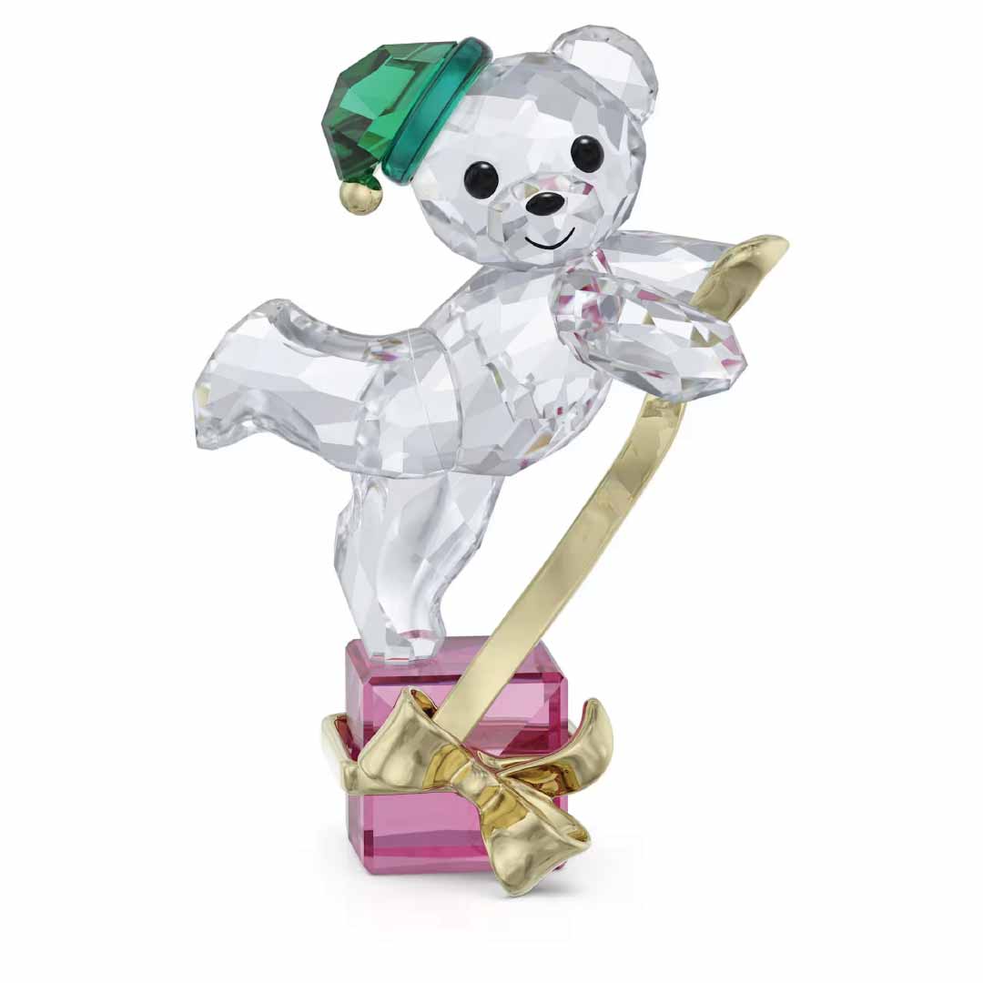 The Swarovski Kris Bear Holiday 2024 Annual Edition Home Décor is a charming collectible that showcases a crystal teddy bear in an endearing pose. It perches on a pink gift box, adorned with a green hat and holding a gold ribbon, while its clear surface beautifully reflects light.