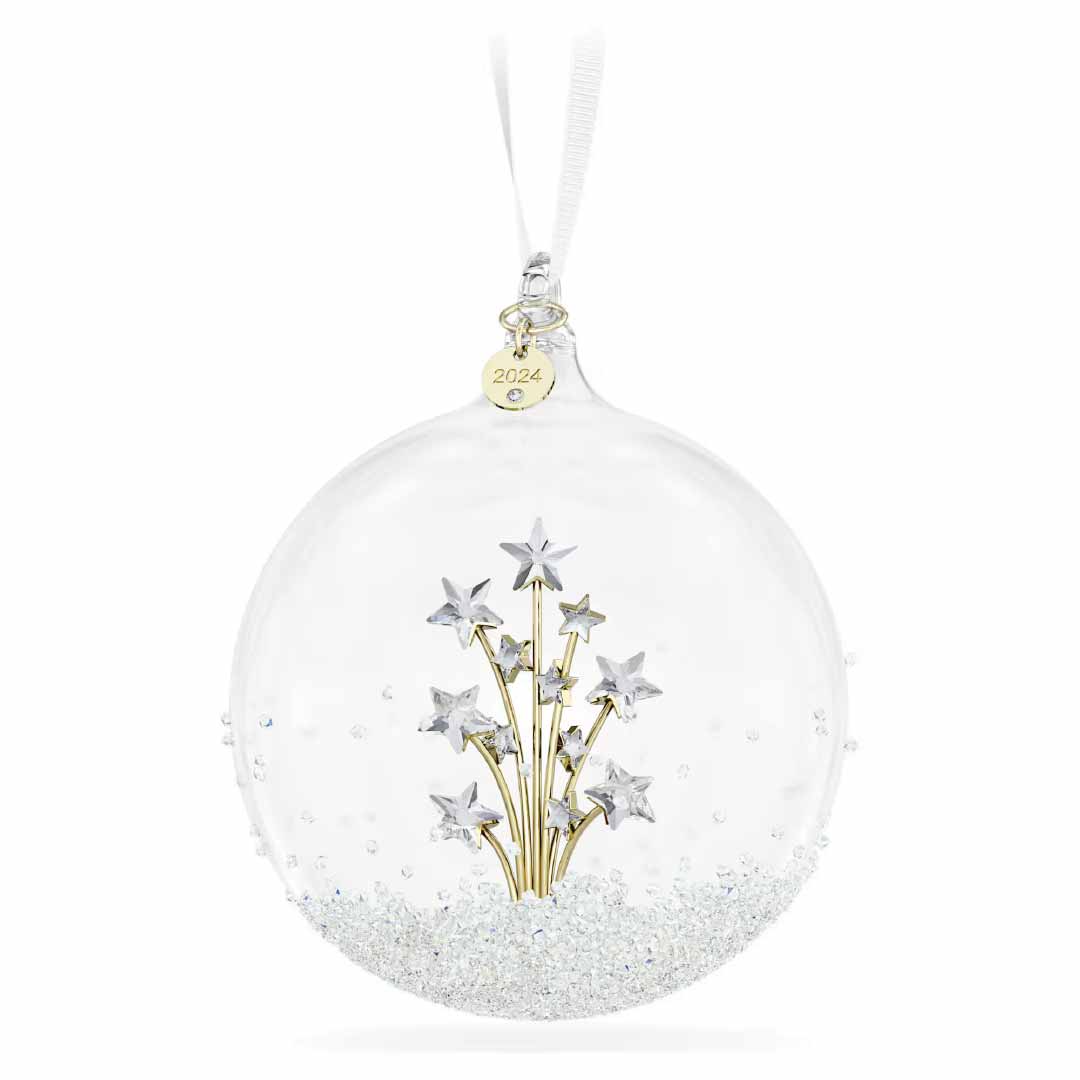 The Swarovski 2024 Annual Edition Ball Ornament is a clear glass piece featuring silver stars on golden stems encased within. It hangs elegantly from a white ribbon, complete with a small gold tag bearing the year "2024." The base is adorned with delicate white and silver glitter, making it a stunning addition to your home décor.