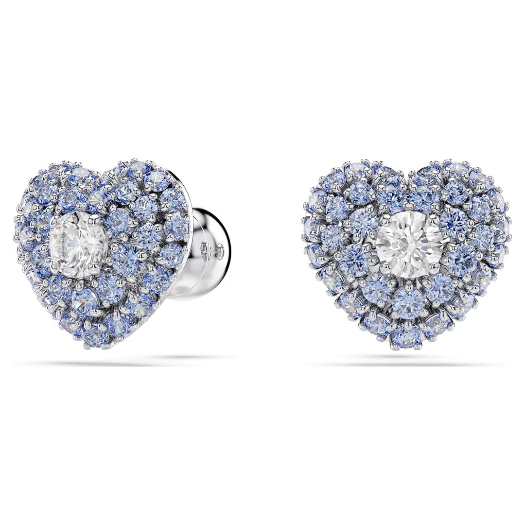 The Swarovski Idyllia Stud Earrings, crafted by Swarovski, feature heart-shaped designs with a central Swarovski Zirconia, encircled by smaller blue diamonds set in silver. These Rhodium-plated studs shine beautifully against a pristine white backdrop.