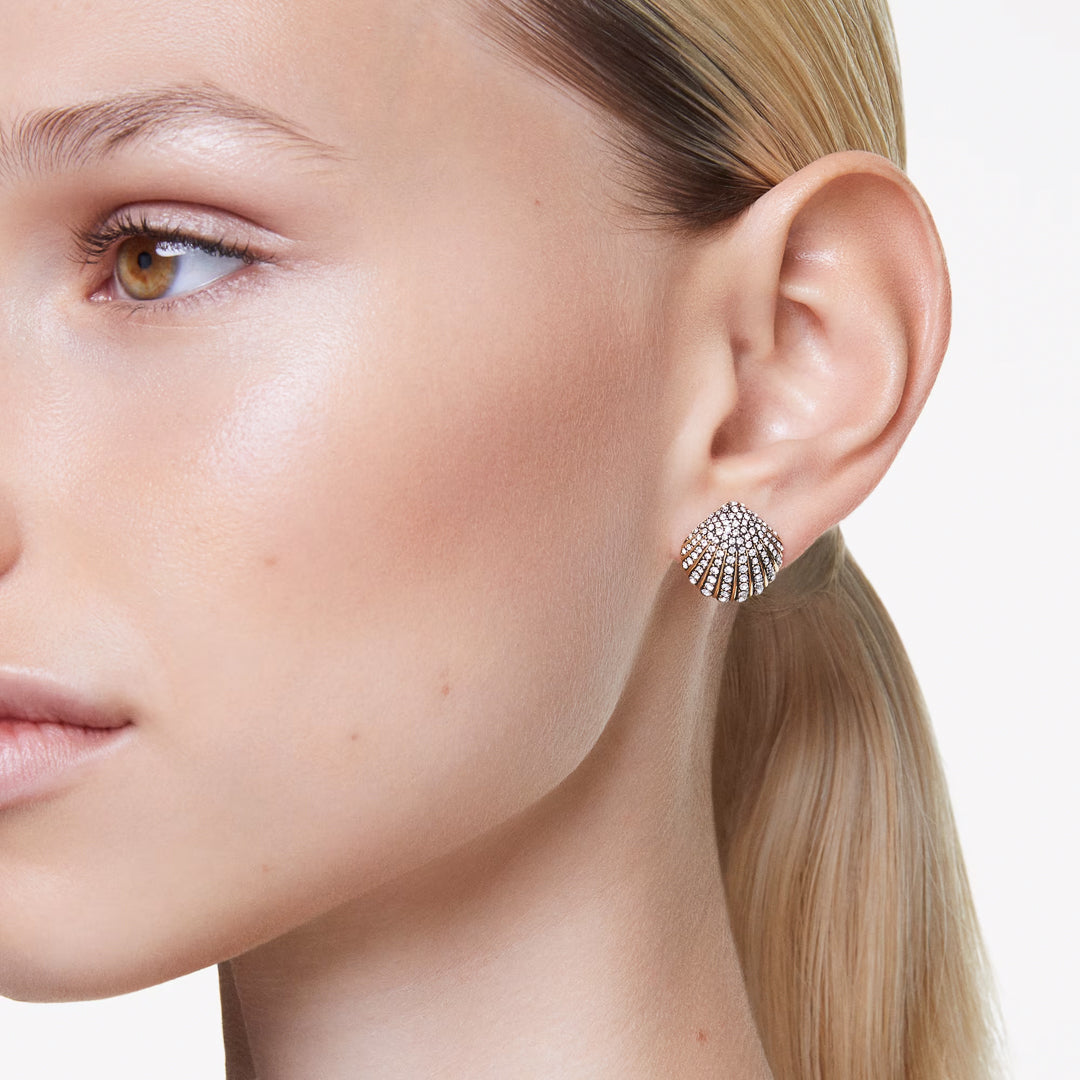 Close-up of a person wearing the elegant Swarovski Idyllia Stud Earrings, featuring a shell motif embellished with crystals. The person's hair is pulled back, highlighting the stunning accessory and their smooth skin against a softly blurred background.
