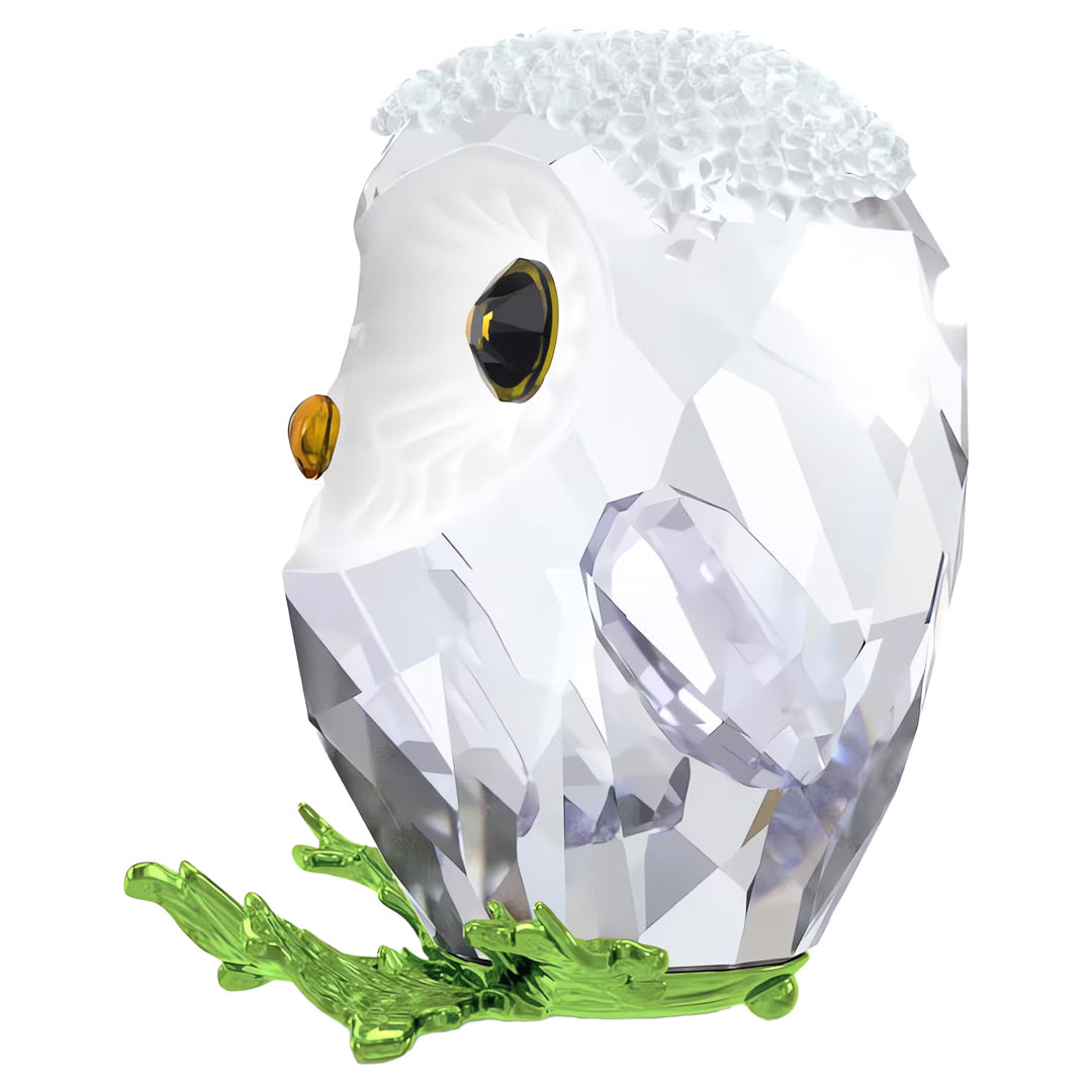 The Swarovski Idyllia Baby Owl Home Décor is an exquisite crystal ornament showcasing a baby owl with a textured surface and frosted white finish, complemented by its striking black eyes and orange beak. The owl sits elegantly on green crystal branches, making it a sophisticated piece that enhances the artistry of any room.