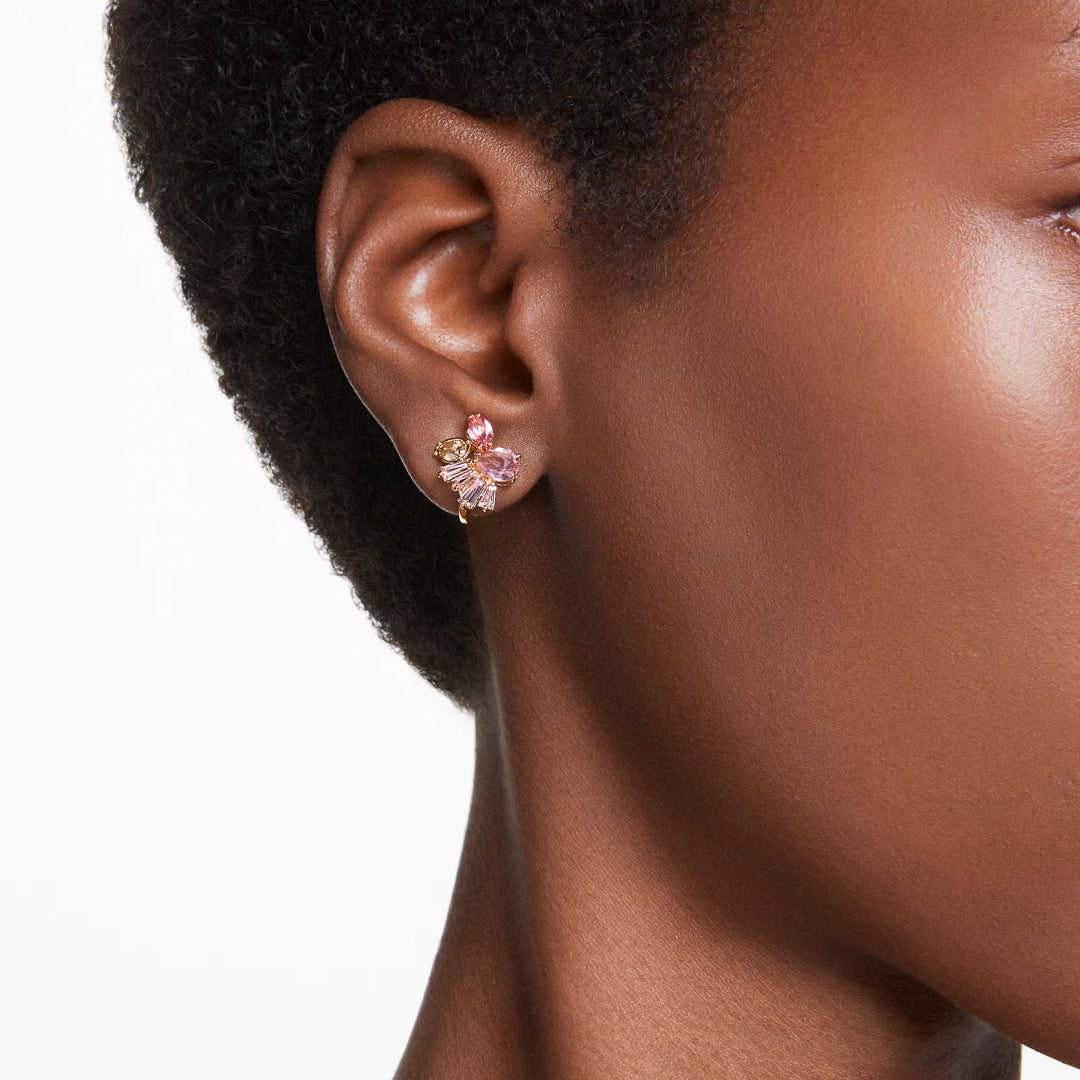 A close-up showcases a person wearing Swarovski Gema Clip Earrings, which are adorned with small pink petals and gold accents shaped like cherry blossoms. These decorative earrings from Swarovski add a touch of elegance to the ear against a plain white background.