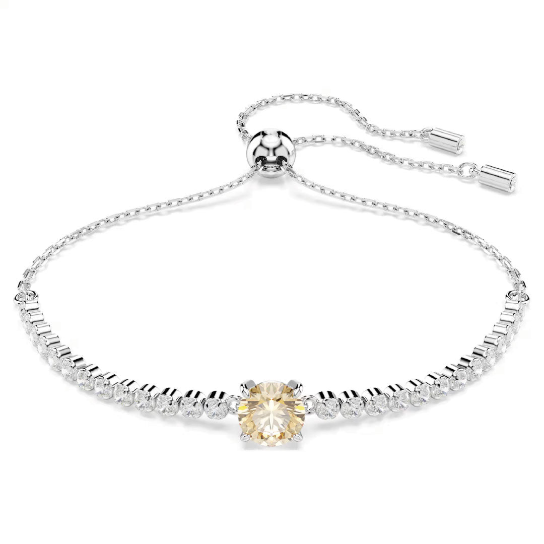 The Swarovski Matrix Tennis Bracelet is a delicate piece, rhodium-plated with an adjustable chain. It showcases a row of small round Swarovski Zirconia and features a striking larger yellow stone in the center.