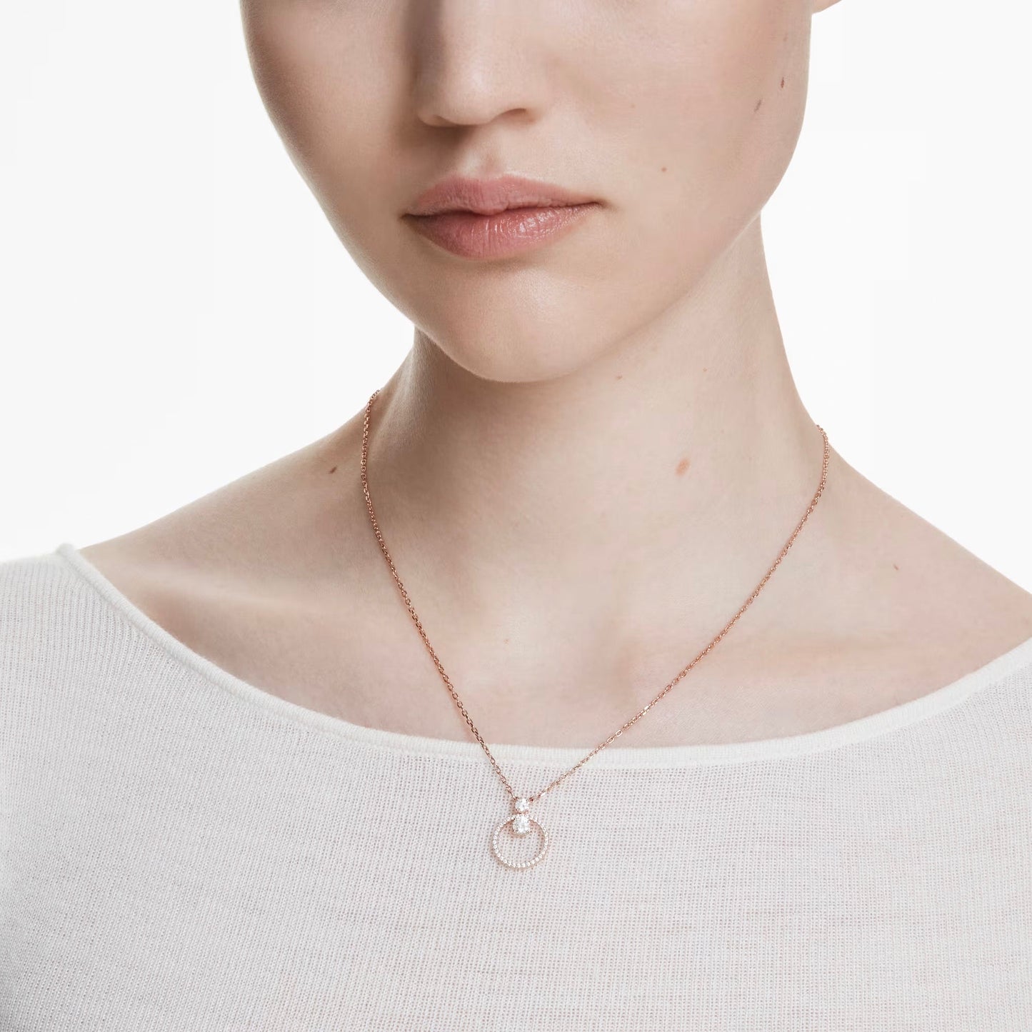 A person dressed in a simple white top is wearing a Swarovski Constella Necklace Pendant, a delicate rose gold-tone plated piece from Swarovski, showcasing an interlocking circle pendant adorned with sparkling Swarovski Zirconia. The background is plain white.