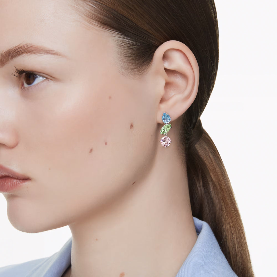 A woman with fair skin and straight brown hair tied back wears Swarovski Gema Multicoloured Drop Earrings. She is wearing a light blue collared shirt and is facing the left side of the image, her earrings shimmering with multicolored crystals.