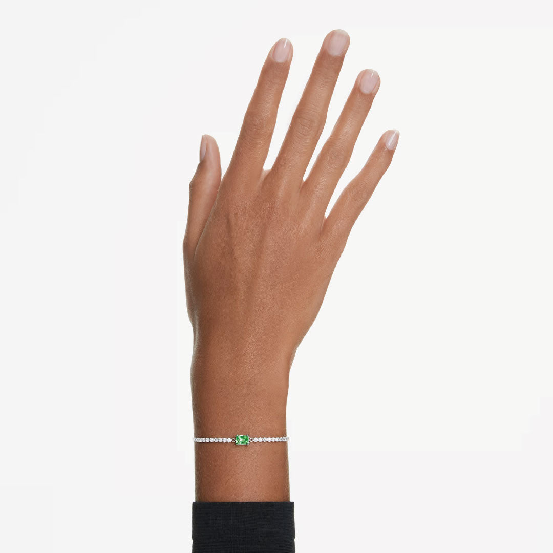 A hand with a black sleeve showcases the Swarovski Matrix Tennis Bracelet, featuring a delicate design with rhodium plating, adorned with a green rectangular stone and small white beads against a pale background.