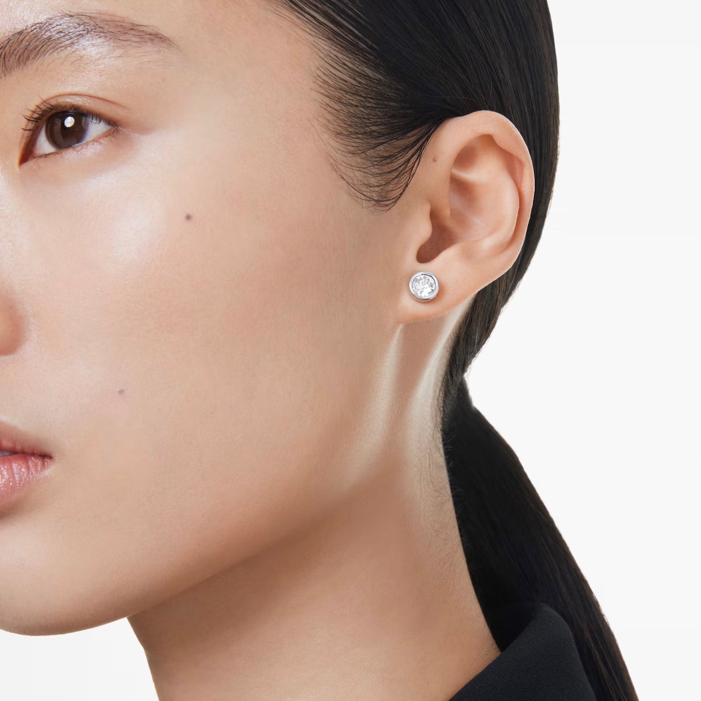 Close-up of a person with straight dark hair pulled back, wearing a single Swarovski Imber Stud Earring. The skin is smooth and glowing, and the plain white background enhances the elegance of the earring from Swarovski.