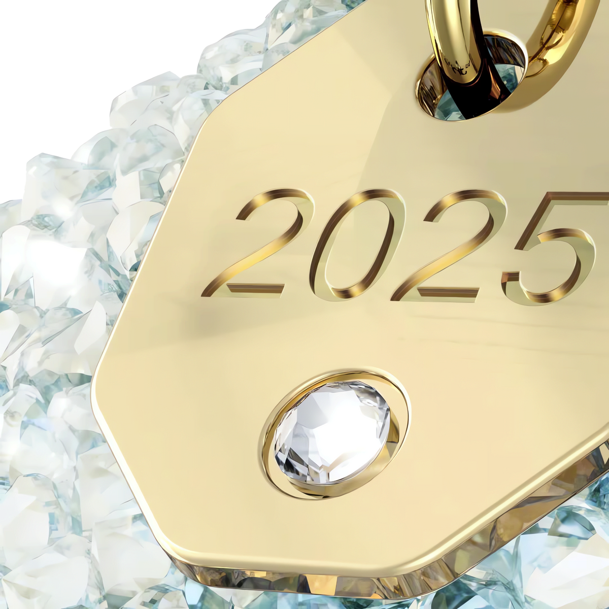 A golden tag engraved with “2025” and featuring a sparkling diamond rests atop a bed of shimmering clear crystals, capturing the elegance of the Swarovski Annual Edition Bell Ornament 2025 by Swarovski—available at Raffi Jewellers, your trusted source for Swarovski Canada treasures.