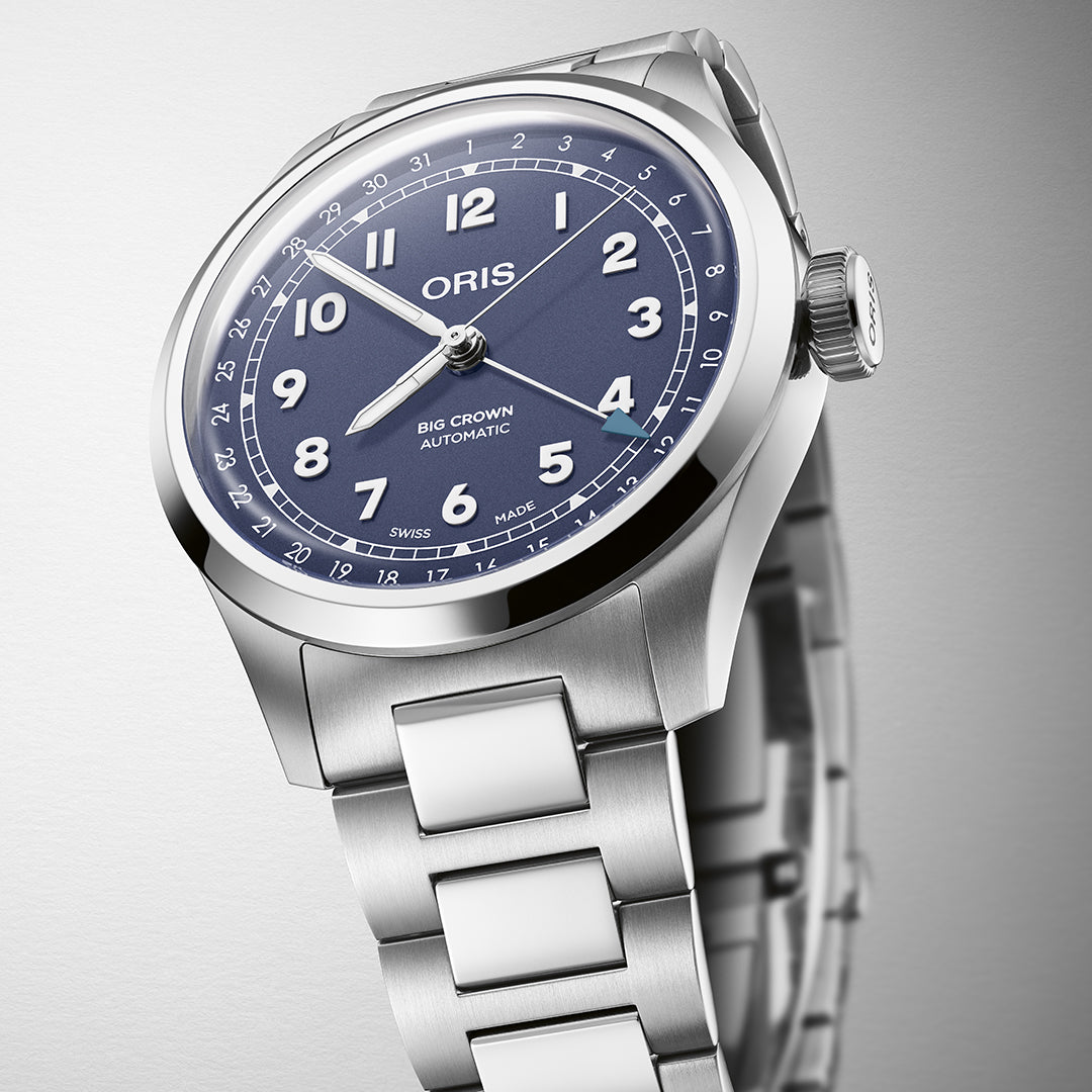 The Oris Big Crown Pointer Date Calibre 754 by ORIS is a silver stainless steel watch with a blue dial, white Arabic numerals, and "Oris Big Crown Automatic" text. This Swiss Made timepiece features automatic mechanical movement, a link bracelet, and a visible crown on the right side against a simple gradient background.