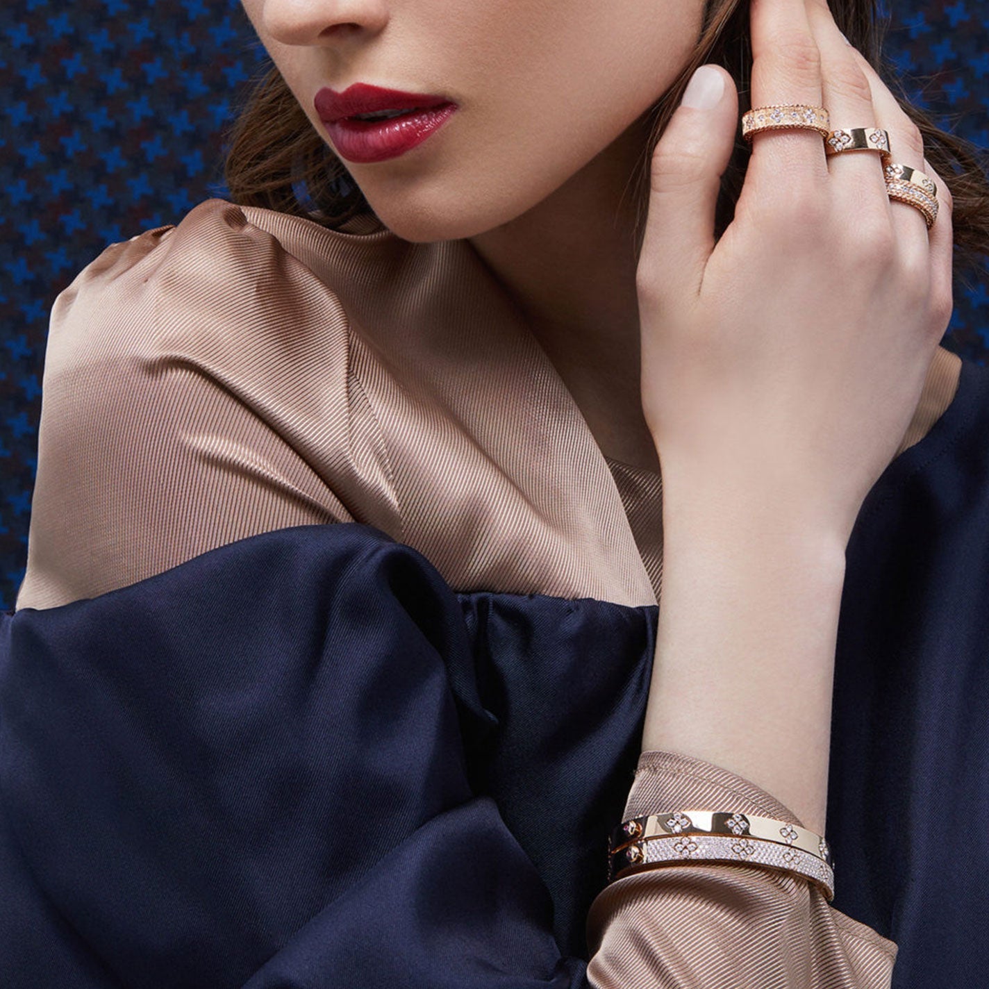 A woman with red lipstick and brown hair wears a satin blouse. Her hands elegantly showcase a stunning Roberto Coin 18K Rose Gold Princess Diamond Satin Ring Band, exuding exquisite design and craftsmanship.