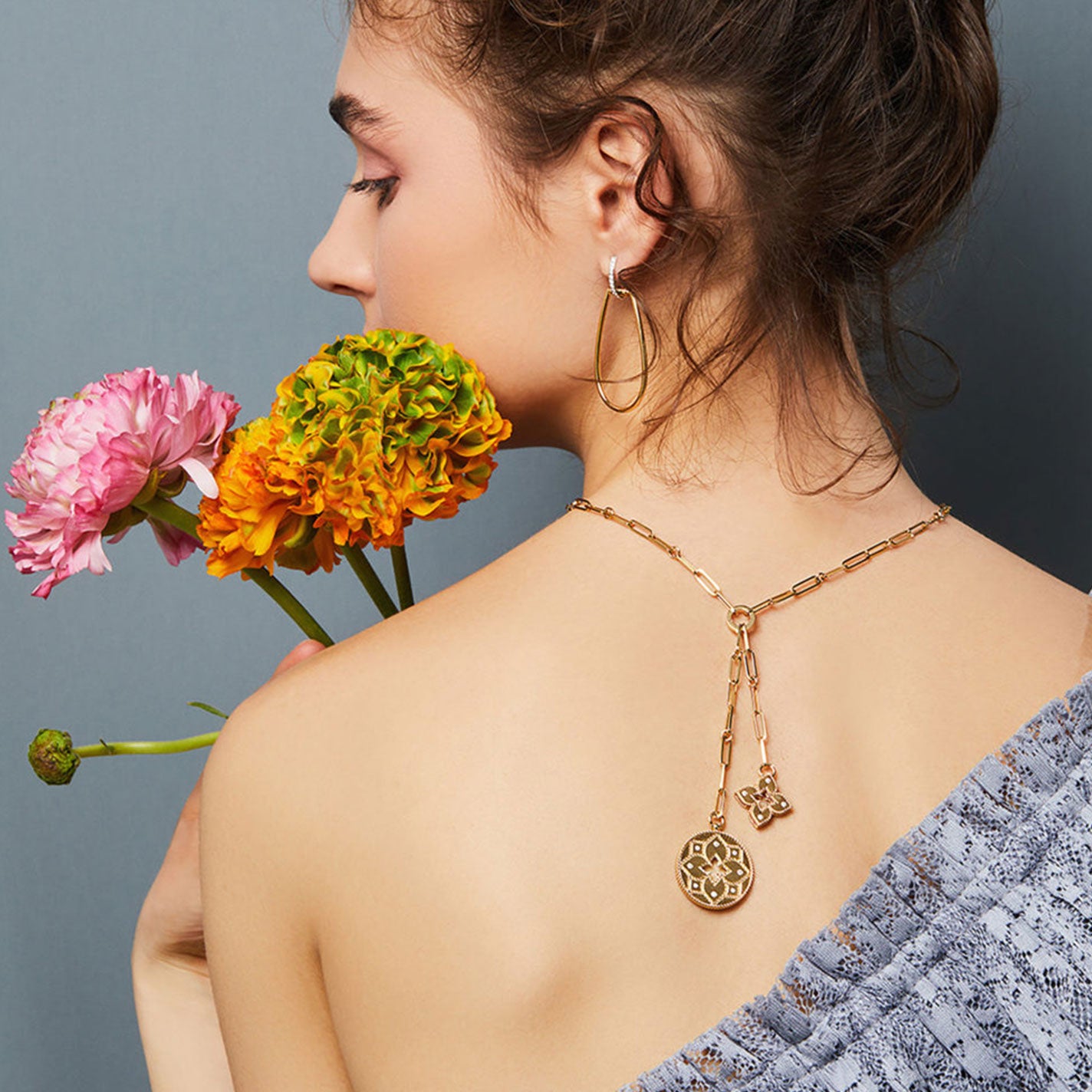 A woman with an updo hairstyle is seen from the back, holding pink and orange flowers. She's wearing a blue lace top and a Roberto Coin Venetian Princess 18K Yellow Gold Diamond Double Medallion Necklace, with the medallions draped elegantly down her back against a gray background.