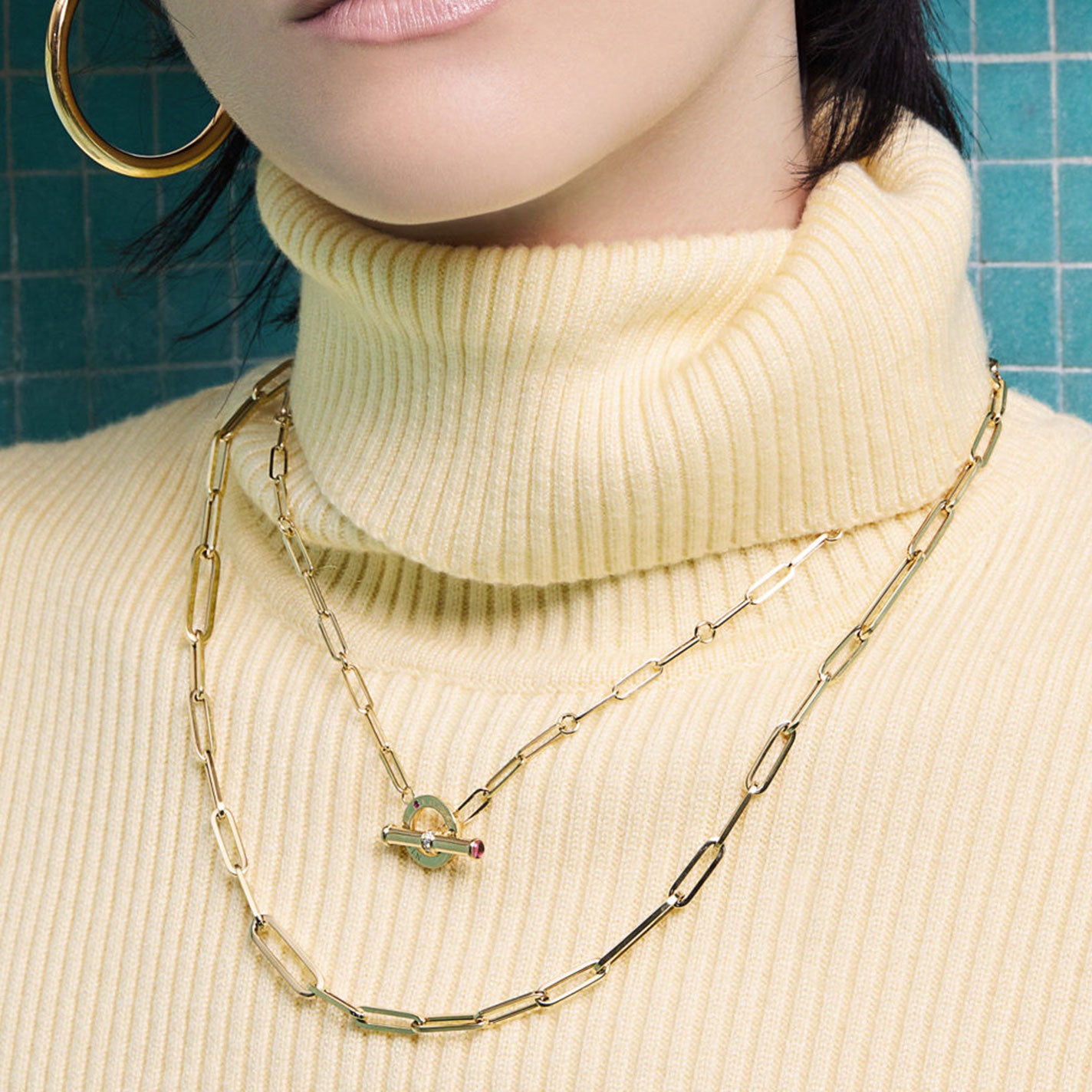 A person wearing a ribbed, cream-colored turtleneck sweater is adorned with two gold chain necklaces. One necklace is the Roberto Coin Designer 18K Yellow Gold Paperclip Chain Necklace, featuring a distinctive diamond toggle closure with a small red gem. The background is a teal, tiled wall.