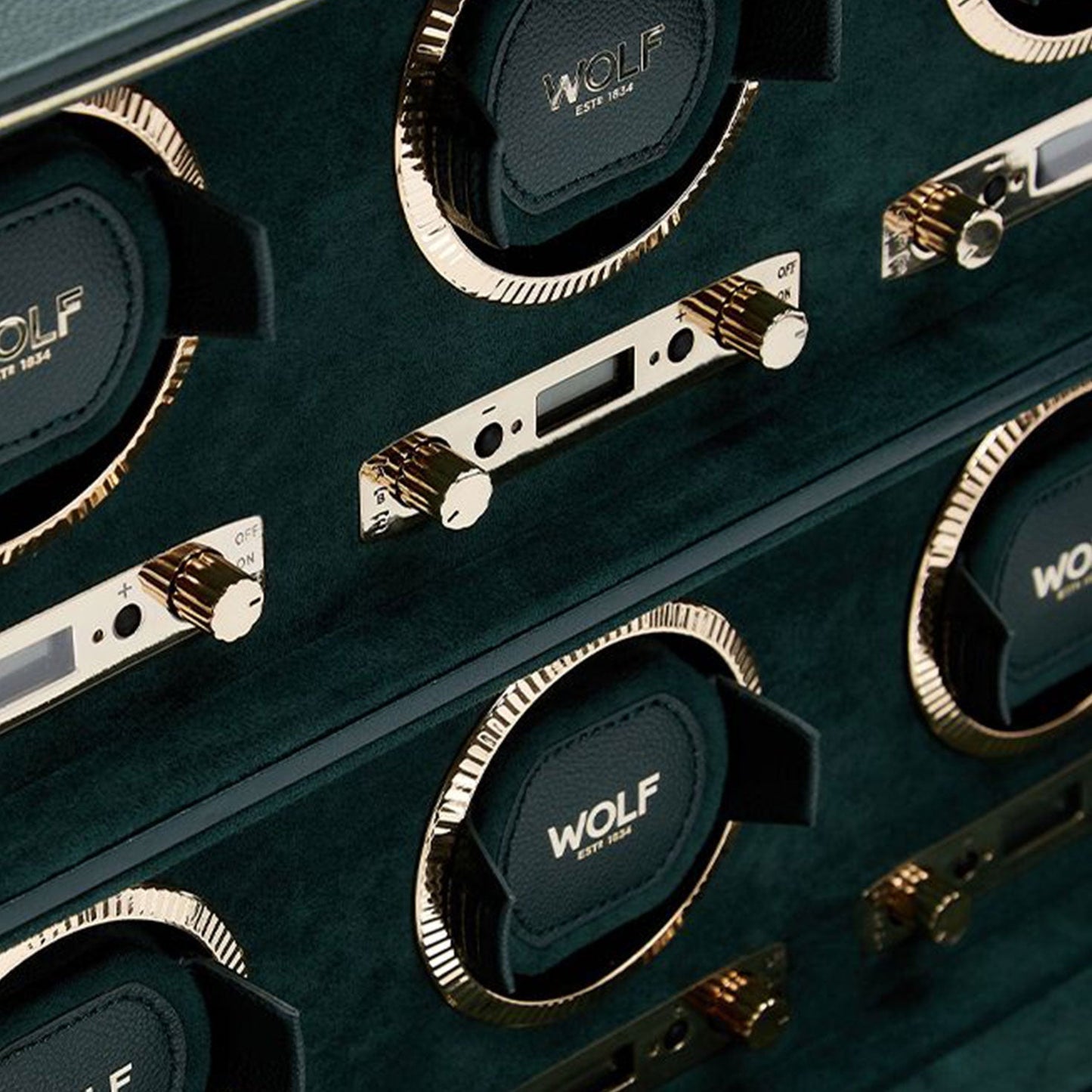 The WOLF 1834 British Racing 6PC Watch Winder features a close-up of its classic luxury display with gold accents. The circular compartments prominently labeled "WOLF" are equipped with control knobs and switches. Set against a dark green background, this design enhances its elegant appearance, reminiscent of British Racing style, emphasizing its timeless sophistication.