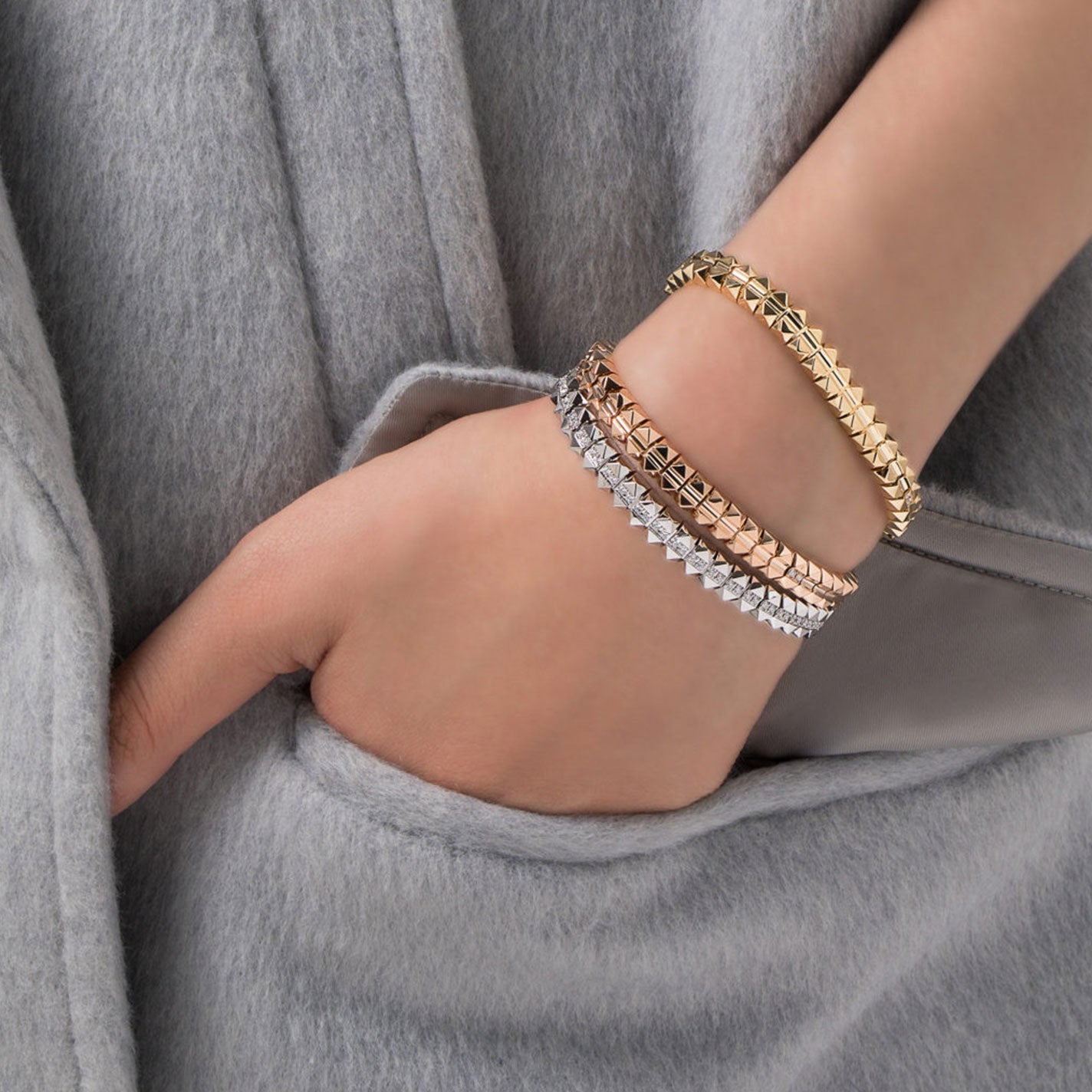 A hand partly tucked in a gray coat pocket, adorned with three pyramid-studded bracelets in gold, silver, and rose gold, catches the light like the Roberto Coin 18K White Gold Diamonds Bangle.