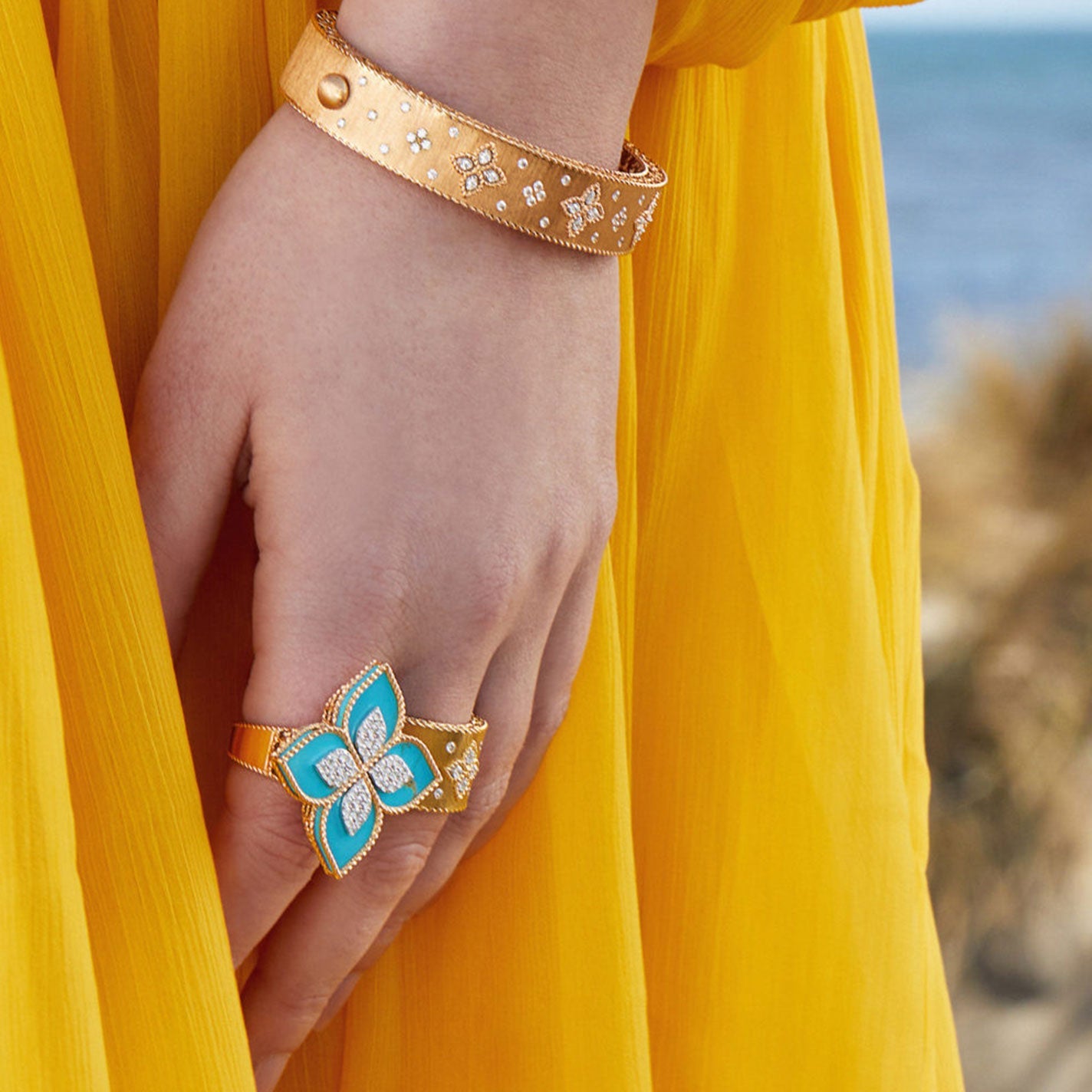 A person in a bright yellow dress showcases a stunning bracelet and the Roberto Coin Venetian Princess 18K Rose Gold Diamond and Turquoise Ring, which is shaped like a butterfly with turquoise wings and diamond accents. The background features a blurred view of a beach and plants.