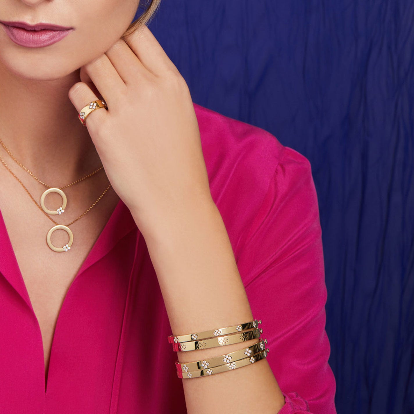 A woman in a vibrant pink shirt accessorizes with a gold ring, layered necklaces, and several gold bangles. Among her jewelry collection is the Roberto Coin Love In Verona 18K White Gold Medium Width Diamond Accent Bangle. The background features a lush blue fabric.