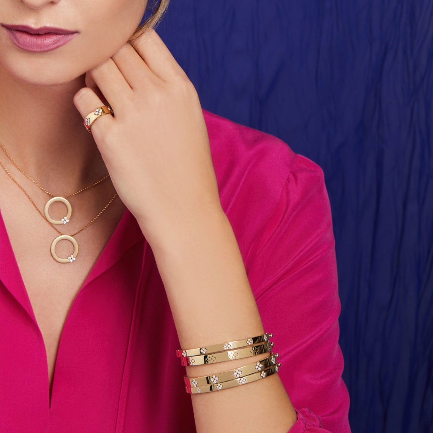 A person in a pink blouse showcases exquisite gold jewelry from Roberto Coin, including three Love In Verona 18K White Gold Diamond Bangles adorned with star motifs, a ring, and two necklaces featuring circular pendants. The collection beautifully stands out against the rich blue fabric backdrop.
