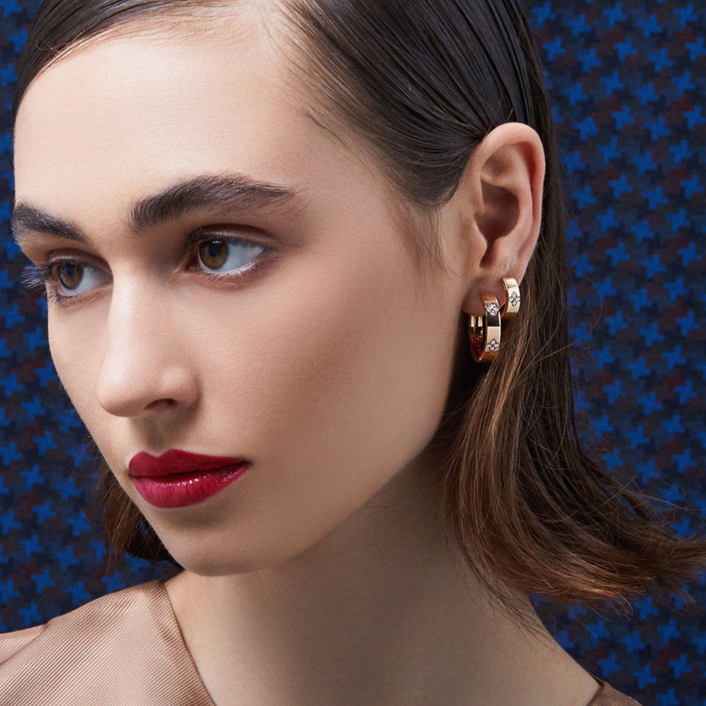 A woman with brown hair and red lipstick gazes to the side, adorning herself with the stunning Roberto Coin Love In Verona 18K White Gold Diamond Accent Small Hoop Earrings. The background showcases a blue pattern with black crosses, echoing the timeless elegance of Love In Verona.