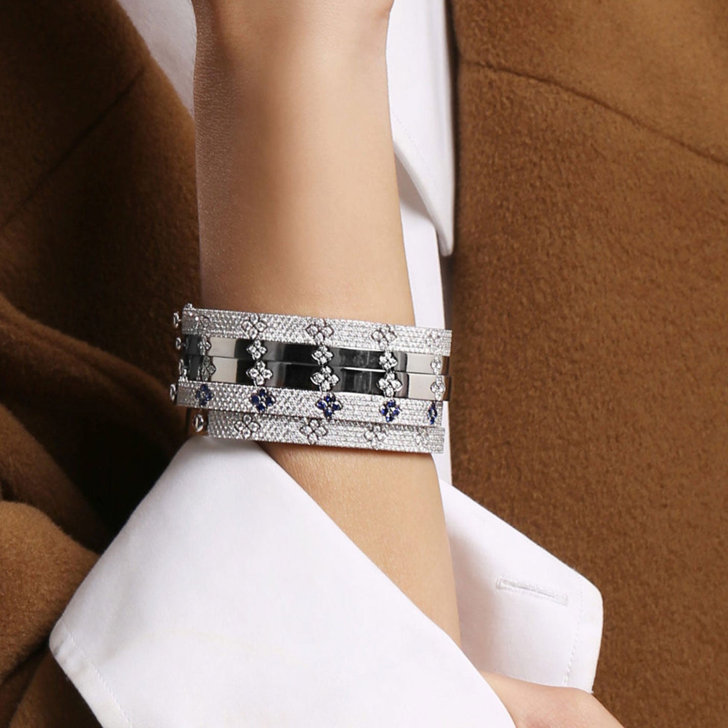 A person wearing the chic Roberto Coin Love In Verona 18K White Gold Pave Diamond Bangle, featuring geometric patterns and small diamond accents, set against the backdrop of a brown coat and a white shirt cuff.