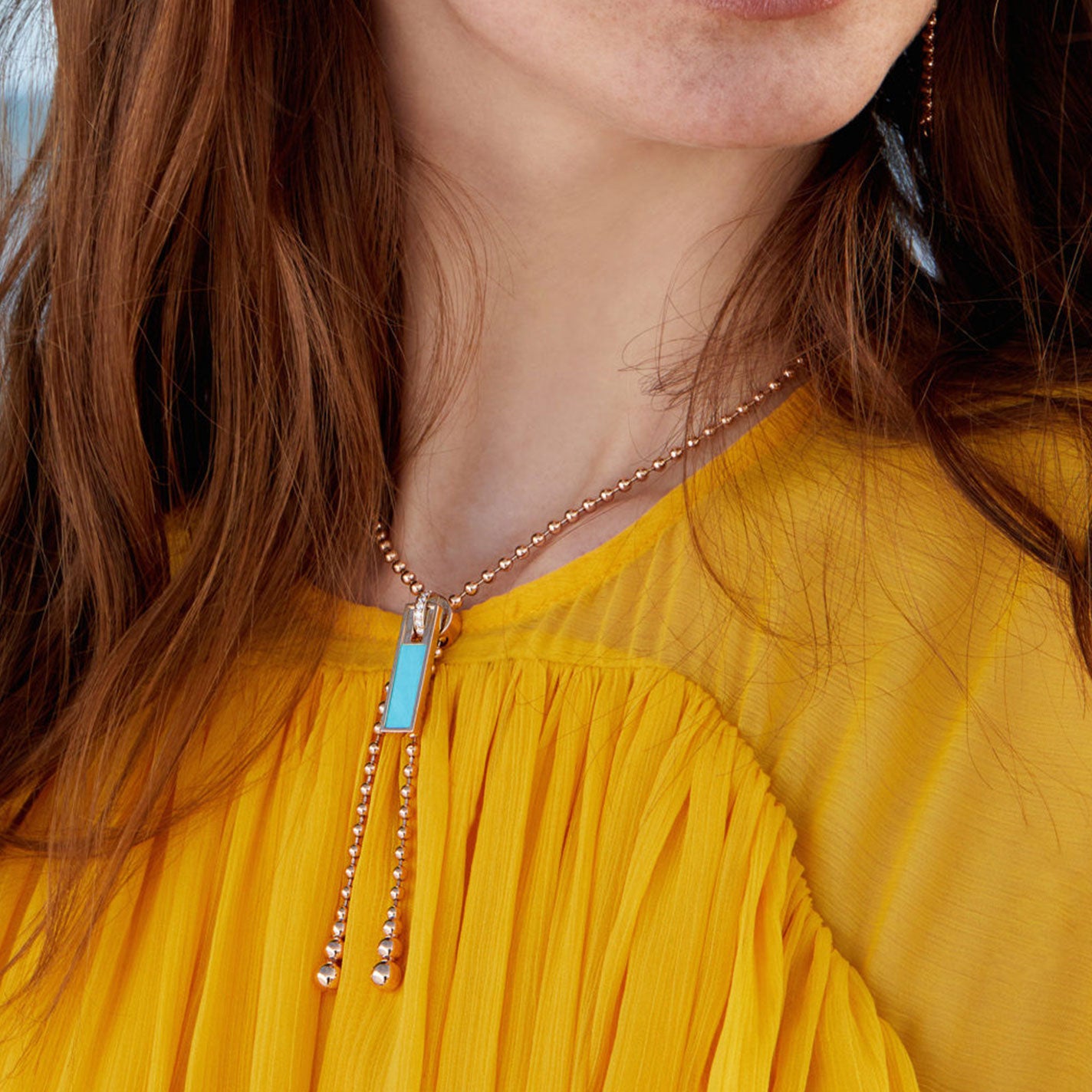 A woman in a mustard yellow top is depicted from the neck down, her cascading long brown hair adding an elegant touch. Adorning her look is the stylish Roberto Coin Art Deco 18K Rose Gold Turquoise and Diamond Zipper Necklace Pendant, which perfectly complements her outfit with its rectangular pendant and beaded chain.