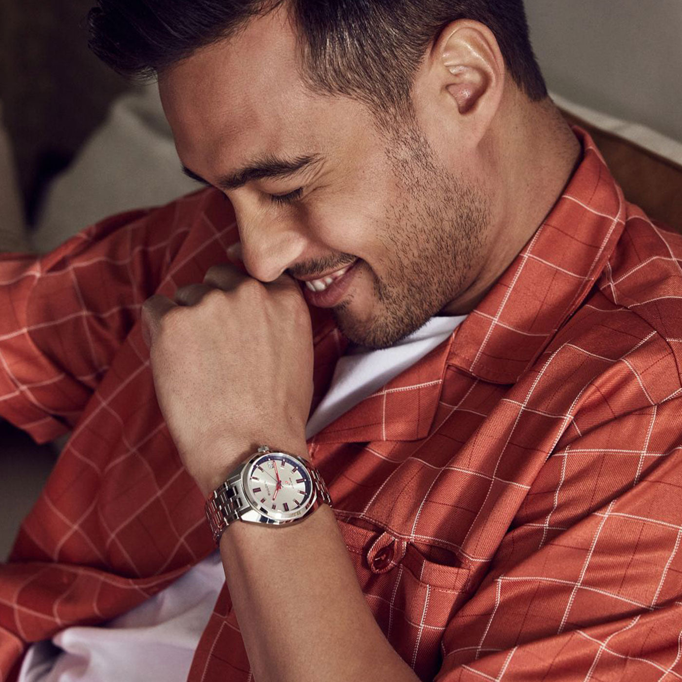 A man in a red checkered shirt smiles as he rests his chin on his hand. Seated comfortably, he wears the Bulova Jet Star NM50 HPQ Precisionist Quartz 40mm watch with its prominent dial, highlighting its precisionist movement. The softly blurred background emphasizes his expression and the limited edition timepiece.