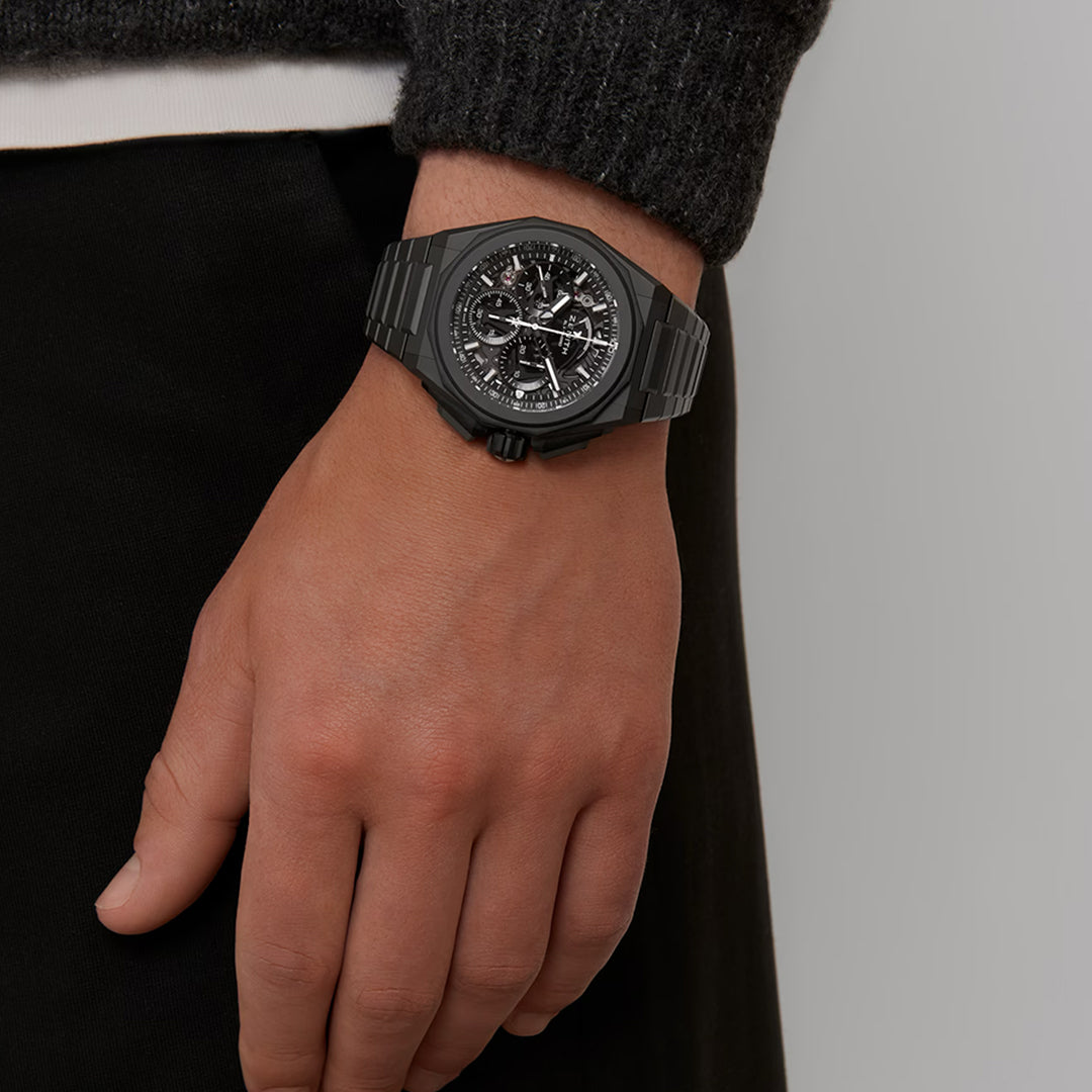 Someone wears a Zenith Defy Extreme El Primero 9004 45mm watch made with precision and detailed geometric patterns in titanium. It complements their dark long-sleeve shirt and black pants while their hand faces downward, showcasing the watch's seamless blend of form and function.