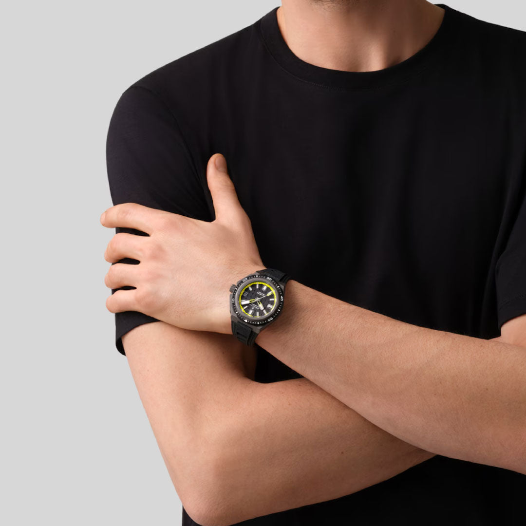 A person in a black t-shirt crosses their arms, showcasing the Zenith Defy Extreme Diver El Primero 3620 43mm Watch with a yellow and black dial and helium escape valve on their left wrist, set against a plain light gray background.