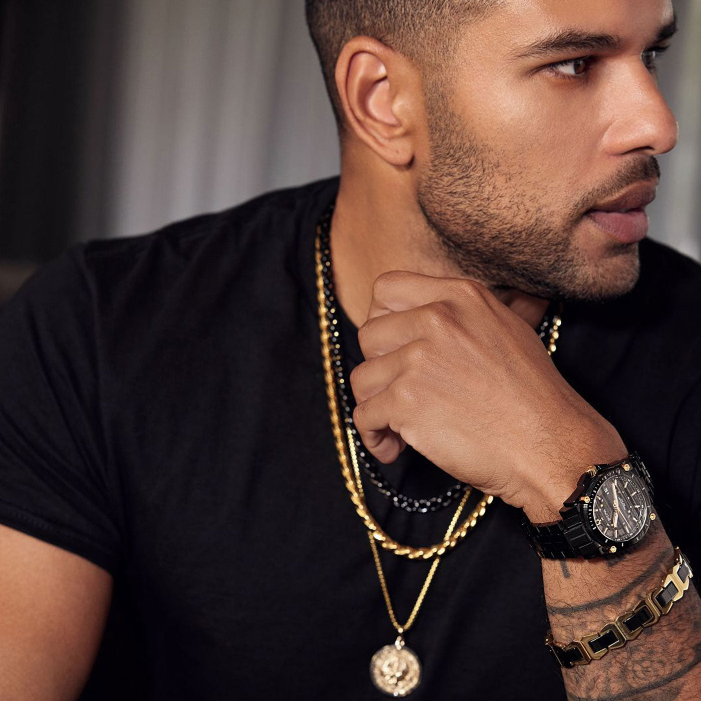 A man with a trimmed beard is wearing a black t-shirt, layered gold necklaces, and the Bulova Icon NM50 HPQ Precisionist Quartz 40mm Watch, part of the men's Icon Collection by Bulova, featuring 316L stainless steel. He's resting his chin on his hand and looking to the side against a blurred background.