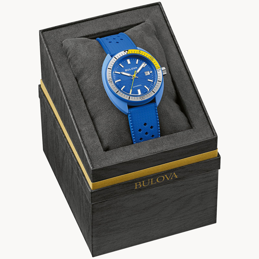 The Bulova Snorkel Blue Tang Quartz 41mm Watch features a vibrant blue face with yellow accents, 100m water resistance, and a perforated strap. It rests on a black cushion inside an elegant black and gold Bulova box.