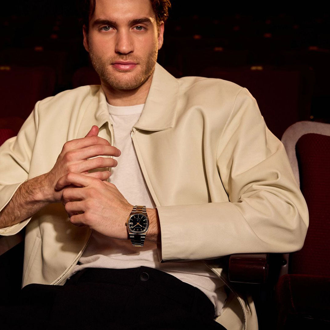 A brown-haired man sits in a theater seat, wearing a cream jacket, white shirt, and black pants. On his left wrist is a Bulova Super Seville HPQ Precisionist Quartz 37.5mm watch. The background shows empty red theater seats.