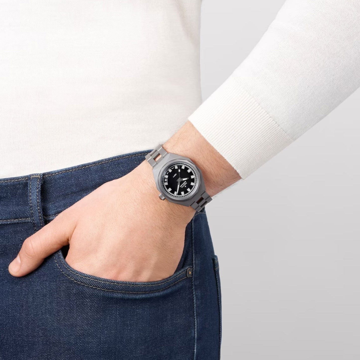 A person dressed in a white long-sleeve shirt and blue jeans casually rests their left hand in their pocket, revealing a Zenith ZENITH Defy Revival Shadow Elite 670 Automatic 37mm watch with a black dial on their wrist, all set against a neutral background.
