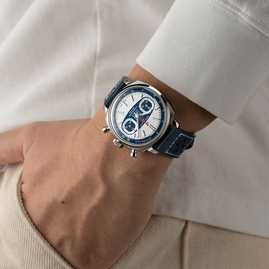 A person in a white long-sleeve shirt and beige pants wears the Breitling Top Time B01 Racing chronograph with a blue strap and white-and-blue dial, their hand casually tucked into their pocket.