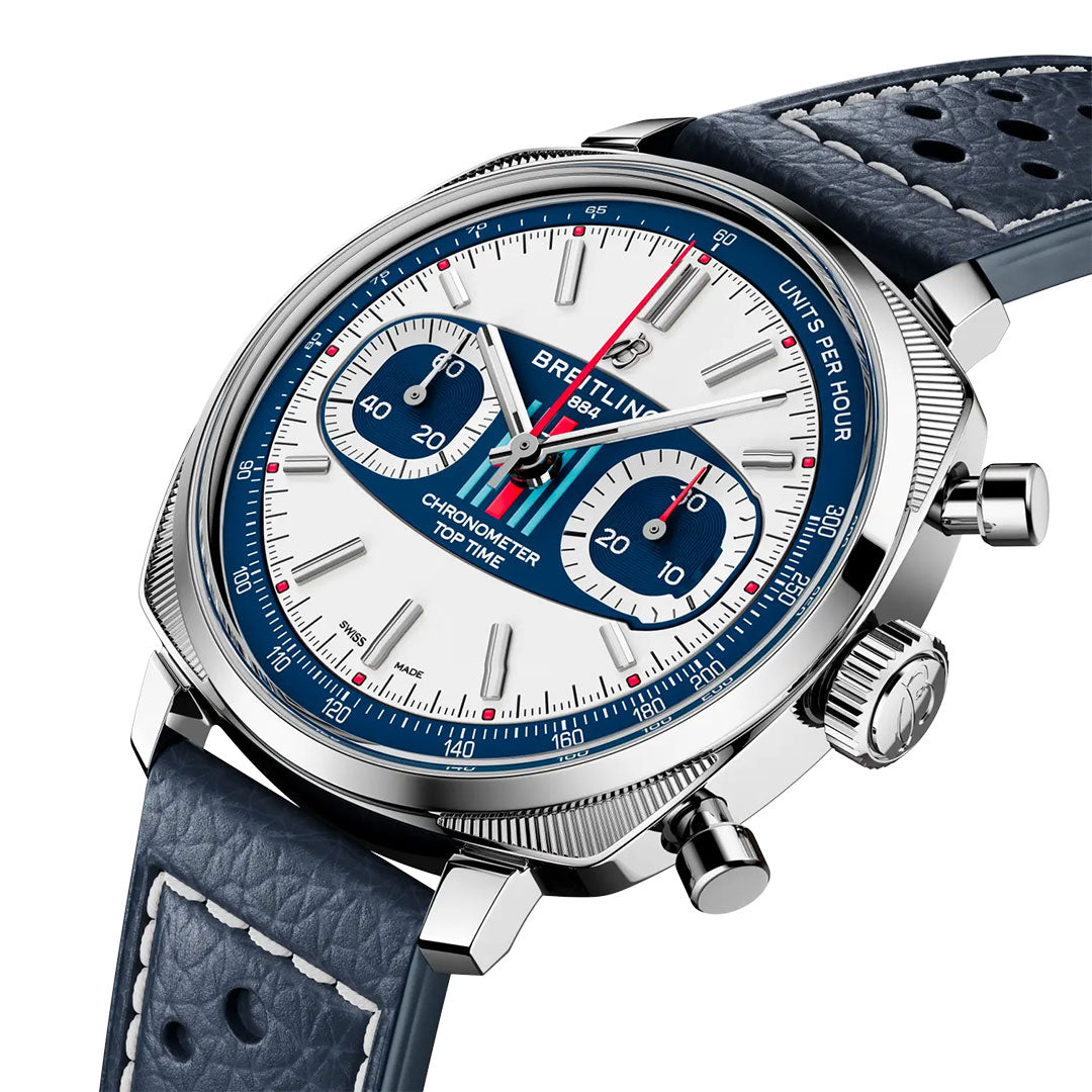 A close-up of the Breitling Top Time B01 Racing chronograph showcases its white and blue dial, three subdials, red accents, and a blue perforated leather strap with white stitching. The stainless steel case reflects classic racing style.