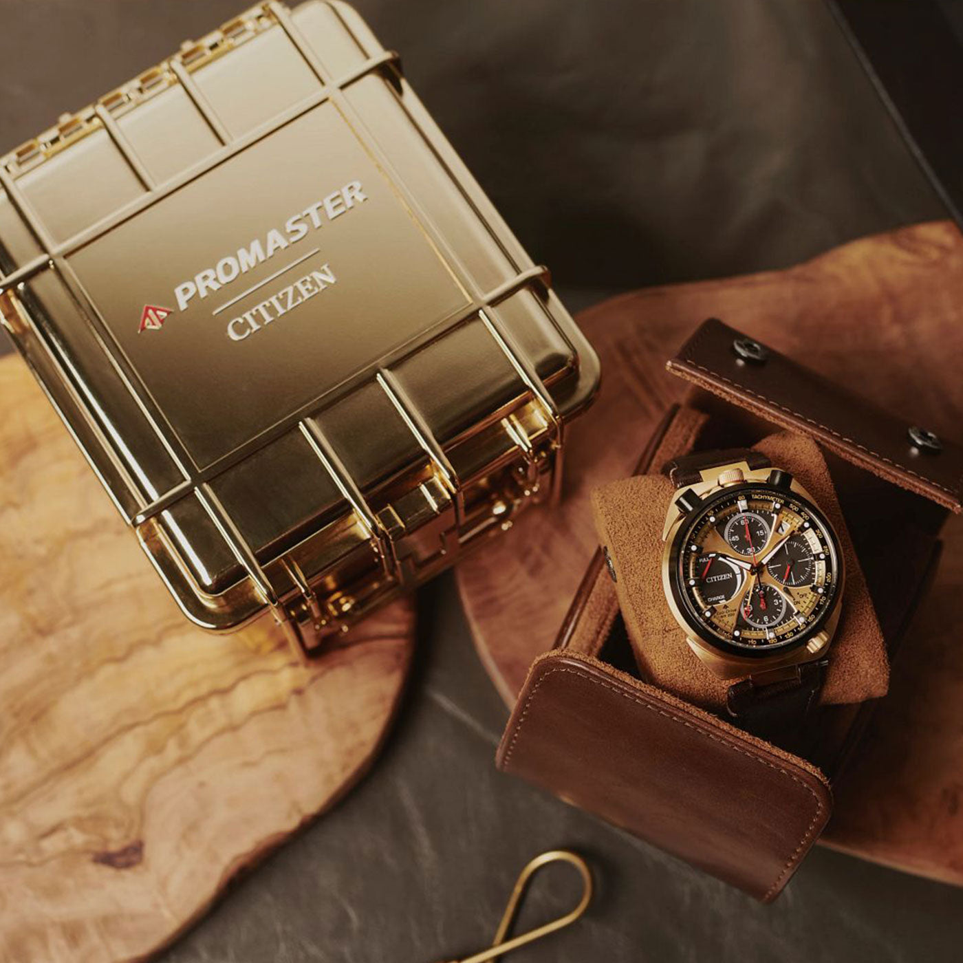 Displayed on a wooden surface is the CITIZEN Promaster Tsuno Chrono Racer E210 Eco-Drive 45mm Watch 50th Anniversary Edition, elegantly housed in a brown leather case. Beside it sits a gold metallic box with the prominent Promaster logo. Completing the setup, a hook tool is subtly placed in the background for an elegant presentation.
