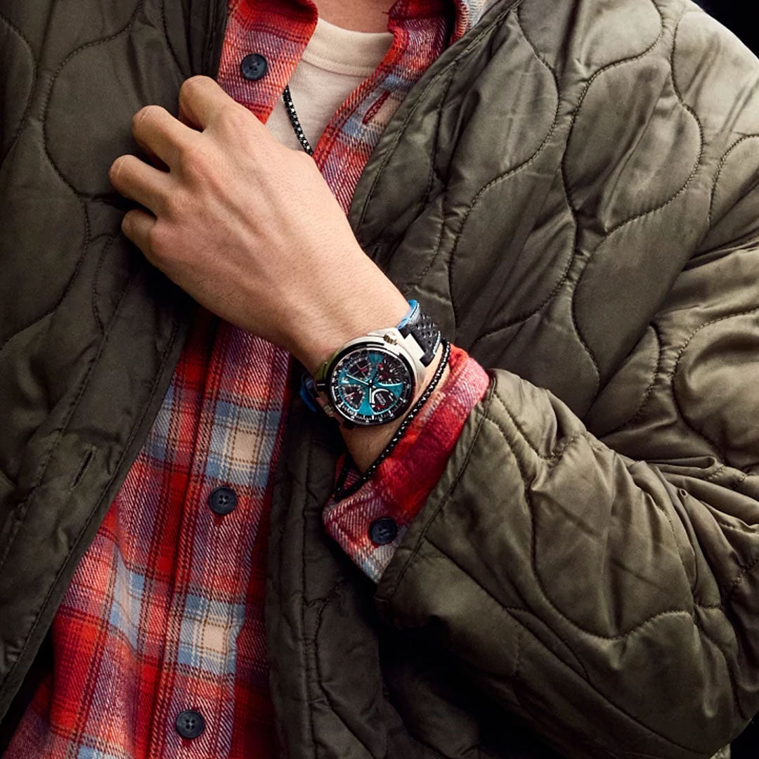Wearing a green quilted jacket over a red plaid shirt, a person shows off their wrist adorned with the CITIZEN Promaster Tsunoc Chrono Racer Limited Edition Eco-Drive 44.5mm Watch, a striking blue and silver chronograph.