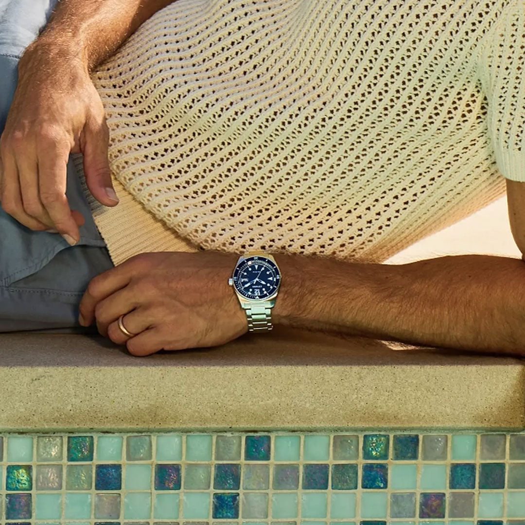 A person relaxes by a pool with blue and green mosaic tiles, dressed in a light knit short-sleeve sweater, gray shorts, and wearing the CITIZEN Terra Force Eco-Drive 43mm Watch by CITIZEN.