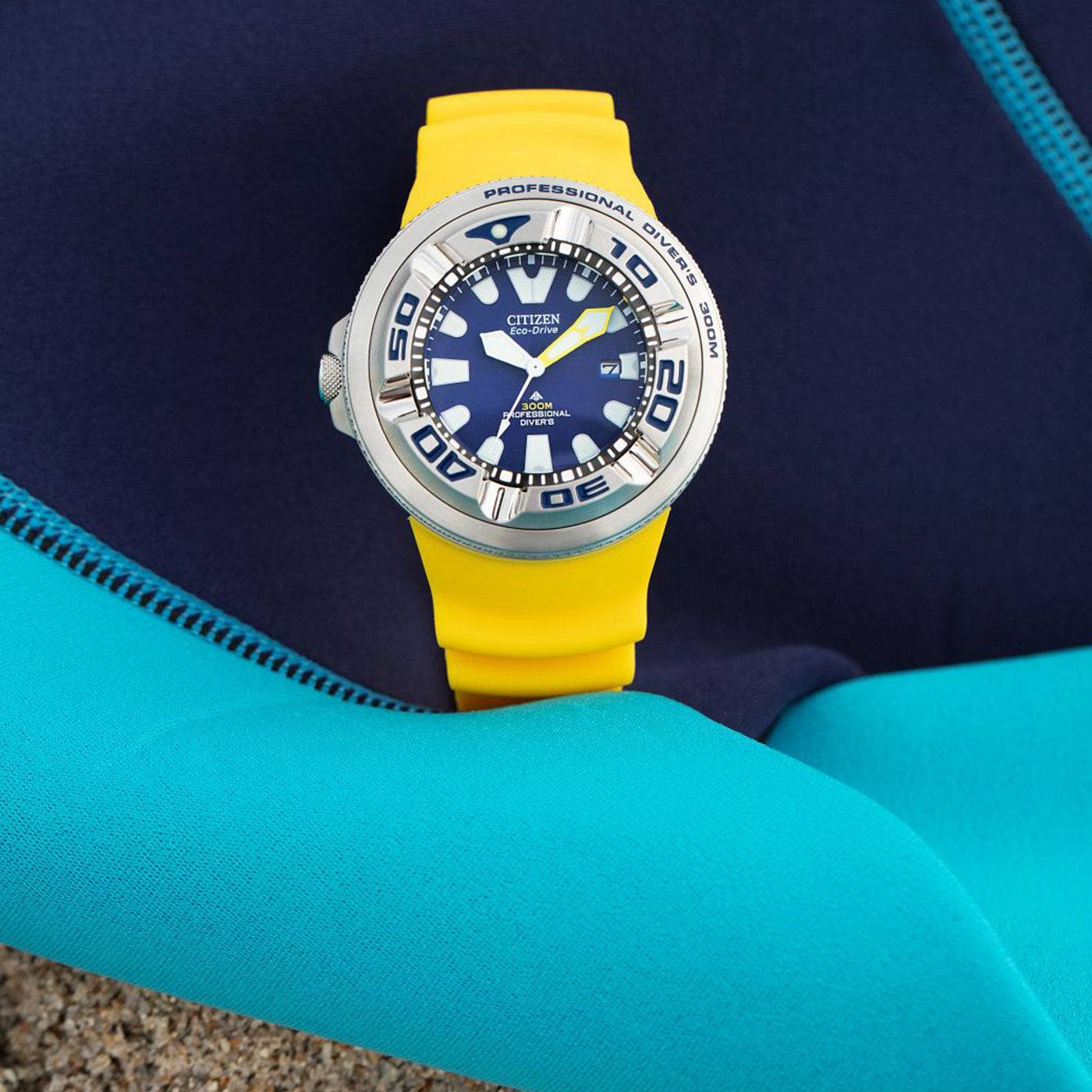 A close-up of the CITIZEN Promaster Dive "Ecozilla" Eco-Drive 48mm Watch, showcasing its striking yellow strap and deep blue face with a date display. The watch rests on a turquoise and navy wetsuit, featuring large silver numbers set against a sandy background, powered by CITIZEN's Eco-Drive technology.