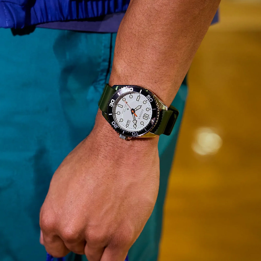 A person wearing a CITIZEN Promaster Dive Eco-Drive 40.6mm watch, featuring a white dial, black bezel, and black strap. The background is blurred with shades of yellow and green.