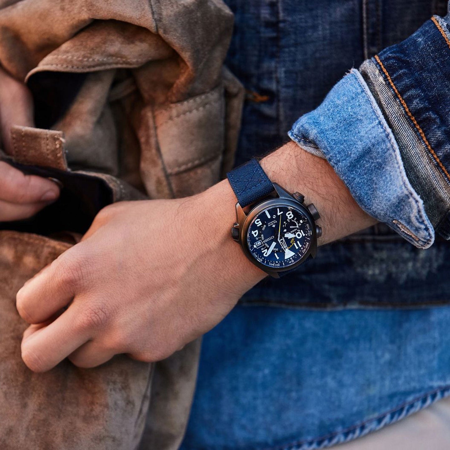 A hand wearing the CITIZEN Promaster Altichron Eco-Drive 47mm Watch, known for its adventure-ready features and intricate dial, reaches into a brown suede bag. The individual, clad in layered denim clothing, achieves a casual and stylish look.