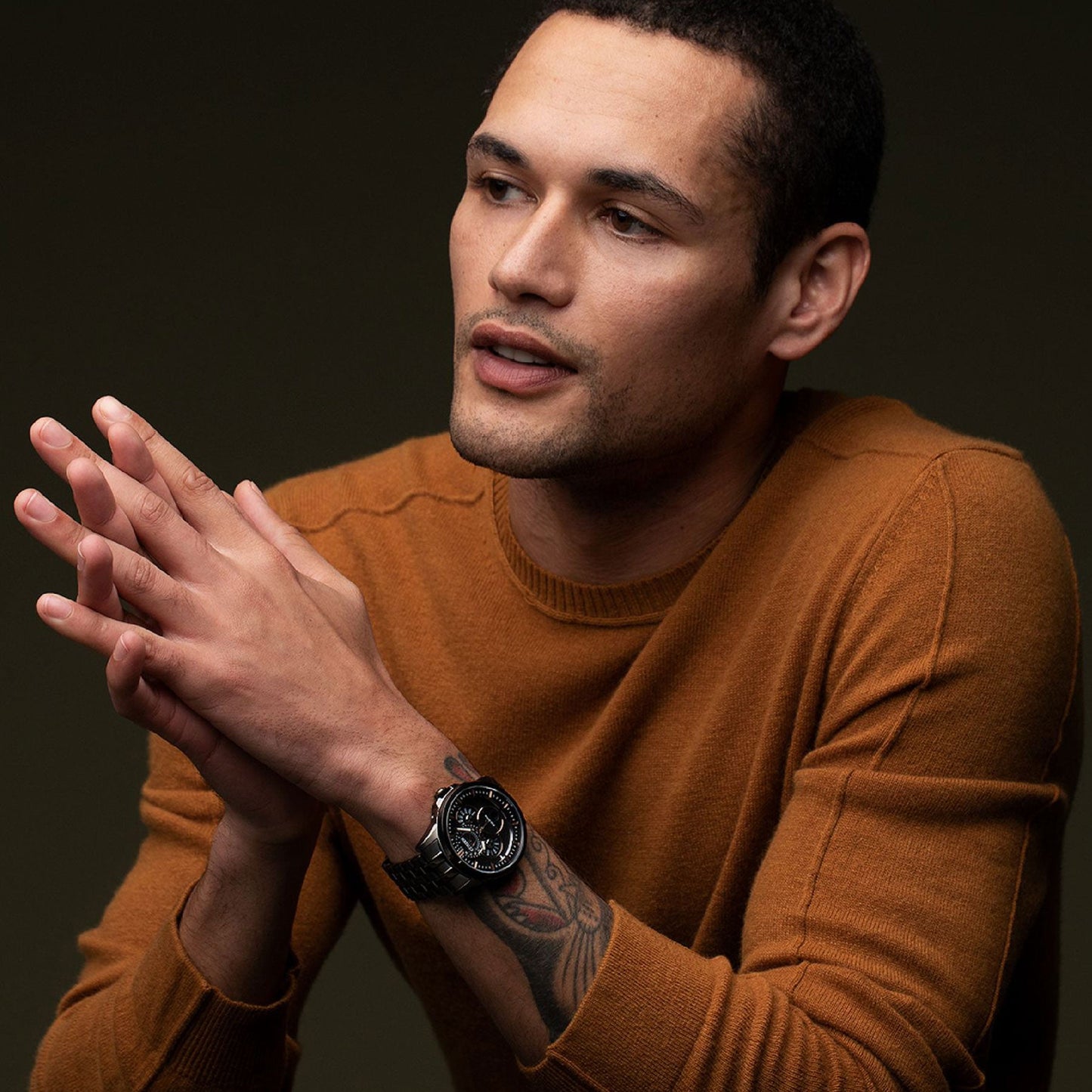 A person wearing a brown sweater and an ion-plated black stainless steel CITIZEN Calendrier Eco-Drive 44mm watch sits quietly with hands clasped, looking to the side against a dark, muted background.