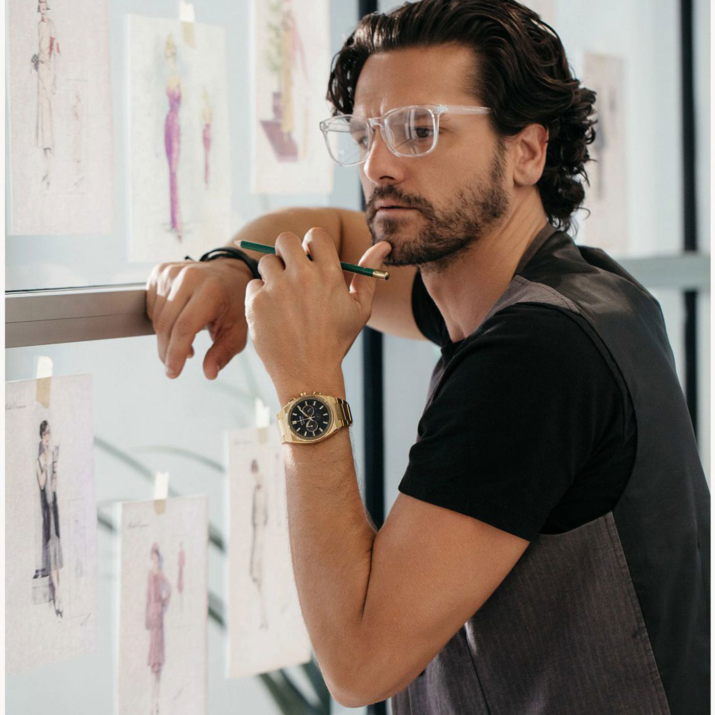 A person with glasses and wavy hair leans against a glass wall, holding a pencil to their mouth. They are wearing a black shirt and the CITIZEN Axiom SC B620 Eco-Drive 43mm Watch, gazing at fashion sketches taped to the wall.