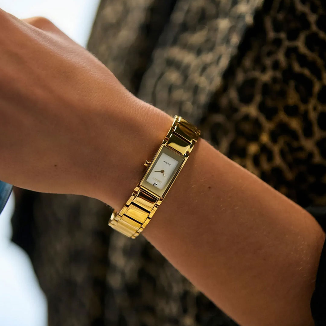 A close-up of a wrist wearing the CITIZEN Axiom Eco-Drive 14.3mm Watch, featuring a rectangular gold-tone case, white dial, and gold band by CITIZEN. The background displays brown and black leopard print fabric.