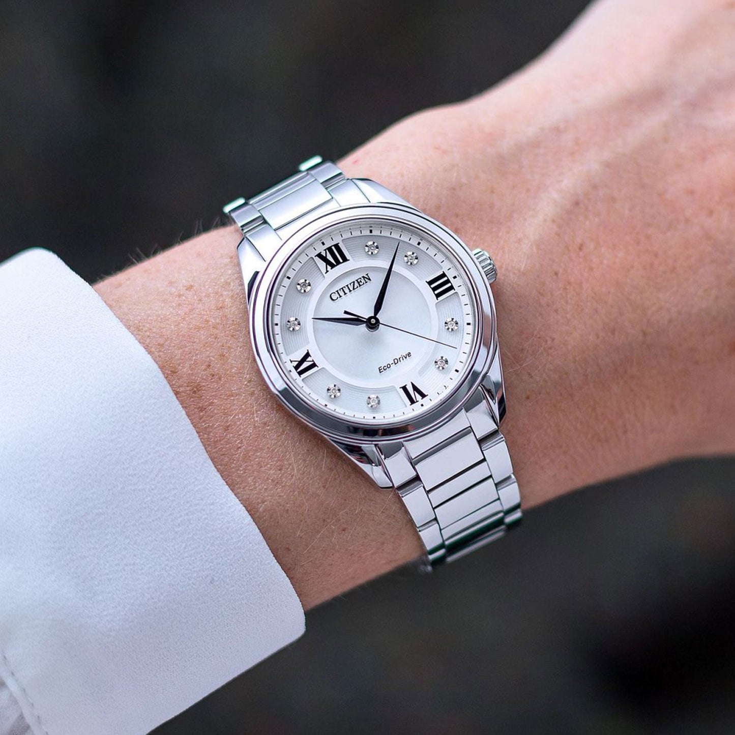 Close-up of a wrist wearing the CITIZEN Fiore Eco-Drive 32mm Watch. This timepiece showcases a white face adorned with Roman numerals and small diamond-like markers, elegantly set in a polished metal band. The wearer complements the watch with a white long-sleeve shirt.