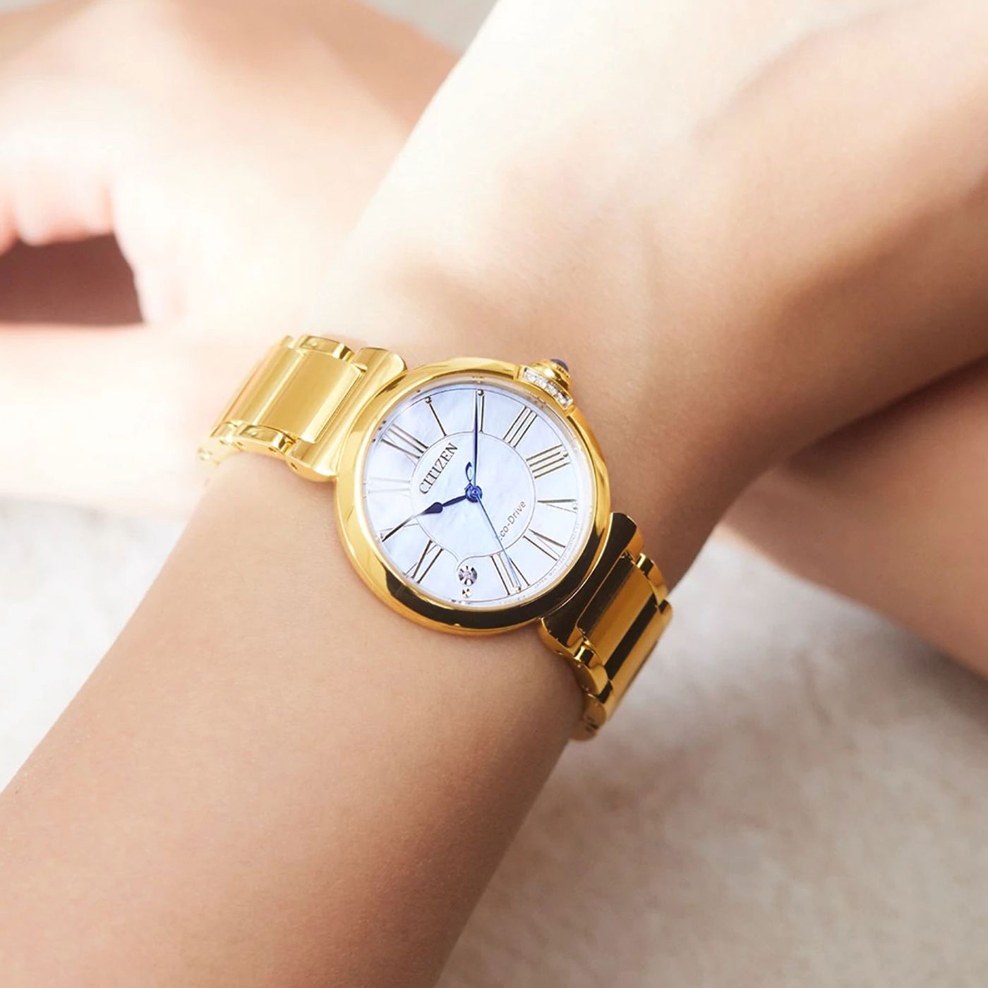 A close-up of a wrist showcasing the CITIZEN L Mae E031 Eco-Drive 29.5mm Watch, adorned with a gold finish, white dial, and black Roman numerals. The metallic strap harmoniously complements its blue hands, while the light background highlights this piece of sustainable luxury.