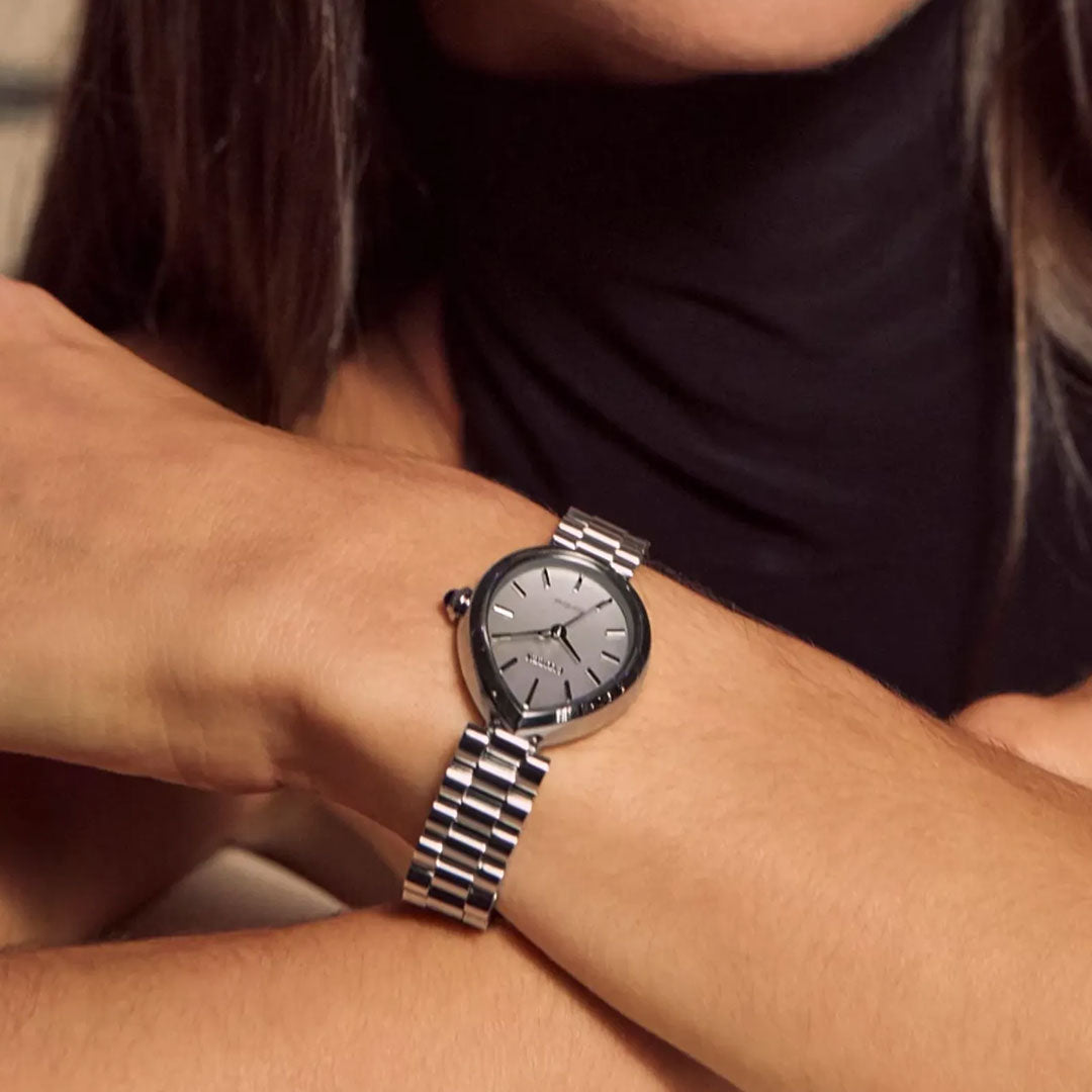 A person in a sleeveless black top wears the CITIZEN L Rainell Eco-Drive 26mm Watch by CITIZEN, featuring a silver-tone stainless steel linked band and round metallic gray face, displaying a time of about 12:48.